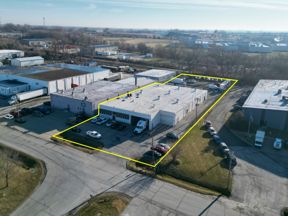 Aerial view of a commercial building with a parking lot, outlined by a yellow boundary, situated in an industrial area with warehouses and open fields in the background.