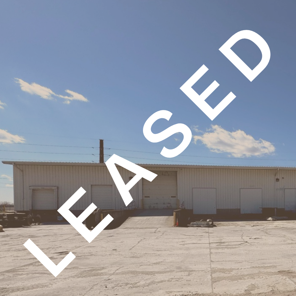A large white industrial warehouse with a loading dock and closed garage doors under a blue sky with clouds.