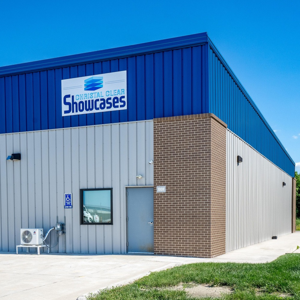 Exterior of a large commercial building with a blue metal roof and gray metal siding, featuring a sign that says 'Christal Clear Showcases.' There is a small window, a door, and some HVAC equipment outside. The building has a brick section on one corner and a sidewalk with grass nearby.