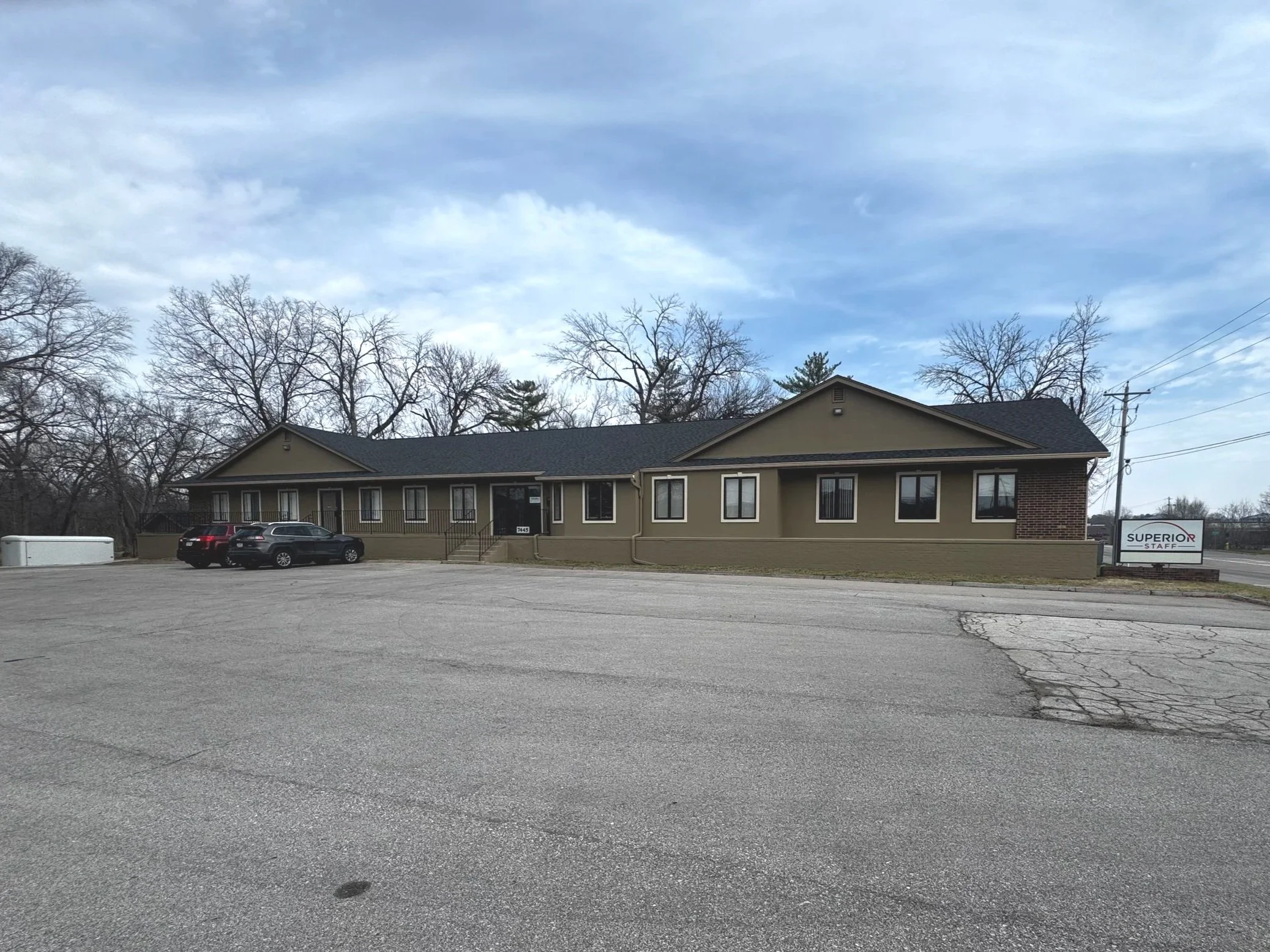 A single-story commercial building with olive green walls, black roof, and a small set of stairs at the entrance. Four cars are parked in front, with a large parking lot and some cracked pavement. There are leafless trees and utility poles in the background under a partly cloudy sky. A sign next to the building reads 'Superior Staff'.