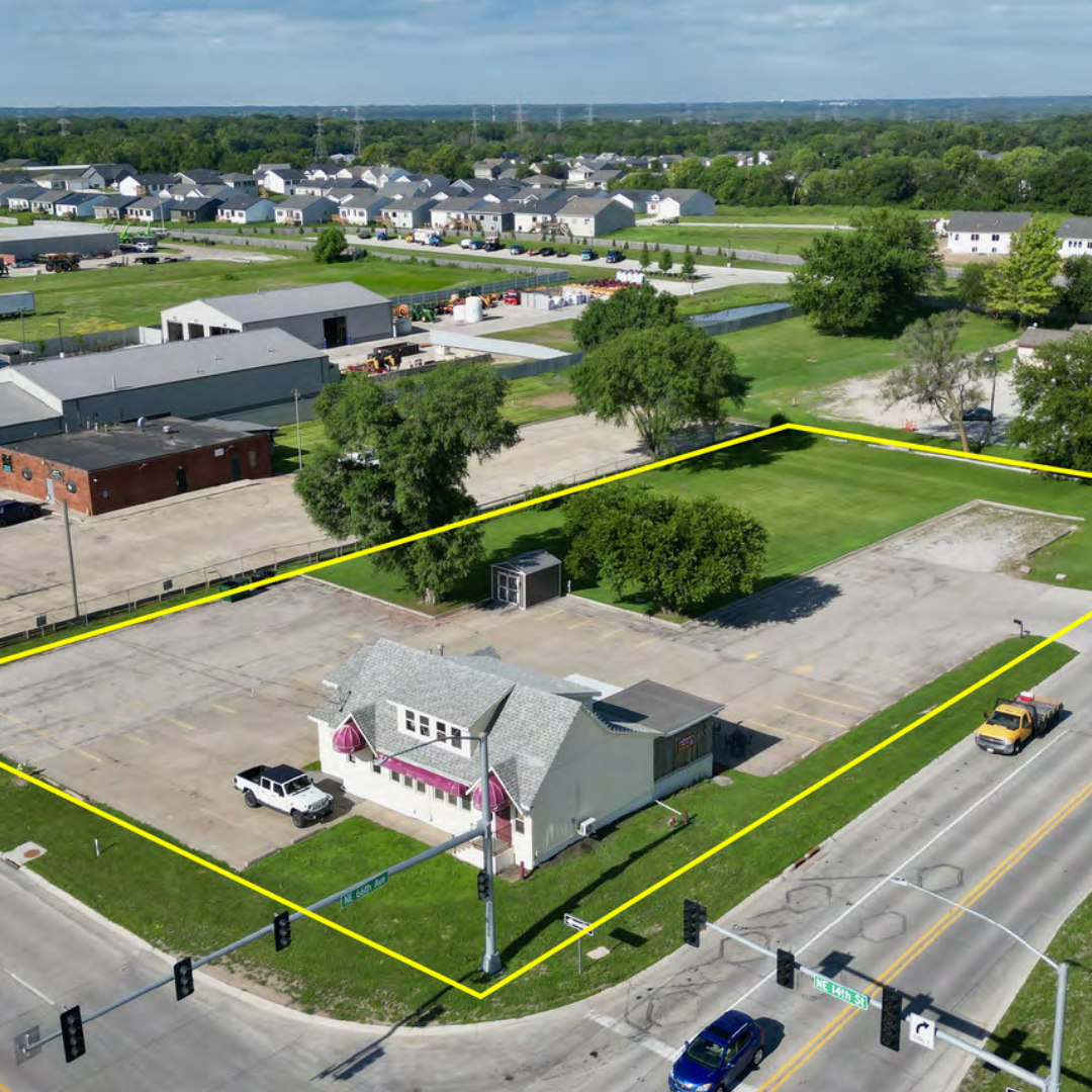 An aerial view of a commercial property outlined with yellow lines, containing a small building with an awning, parking spaces, and trees, situated at an intersection with traffic lights.