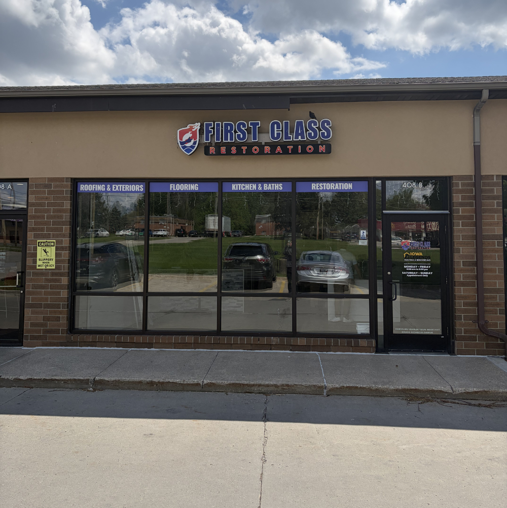  storefront with a sign that reads "First Class Restoration," with additional signage indicating services like roofing, exteriors, flooring, kitchen & baths, and restoration. There are parked cars visible through the windows and a caution sign on the left side of the storefront.