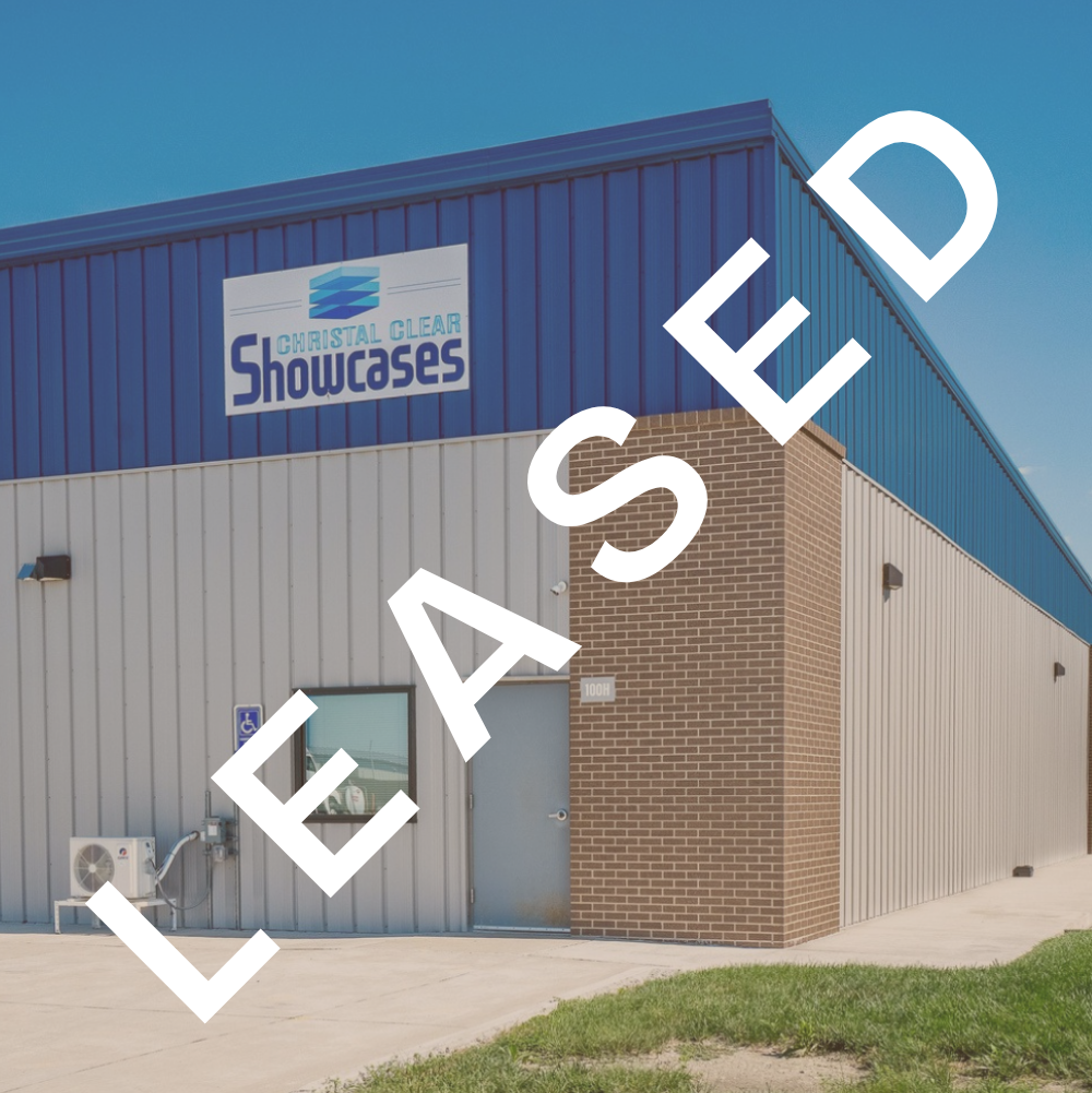 Exterior of a large commercial building with a blue metal roof and gray metal siding, featuring a sign that says 'Christal Clear Showcases.' There is a small window, a door, and some HVAC equipment outside. The building has a brick section on one corner and a sidewalk with grass nearby.