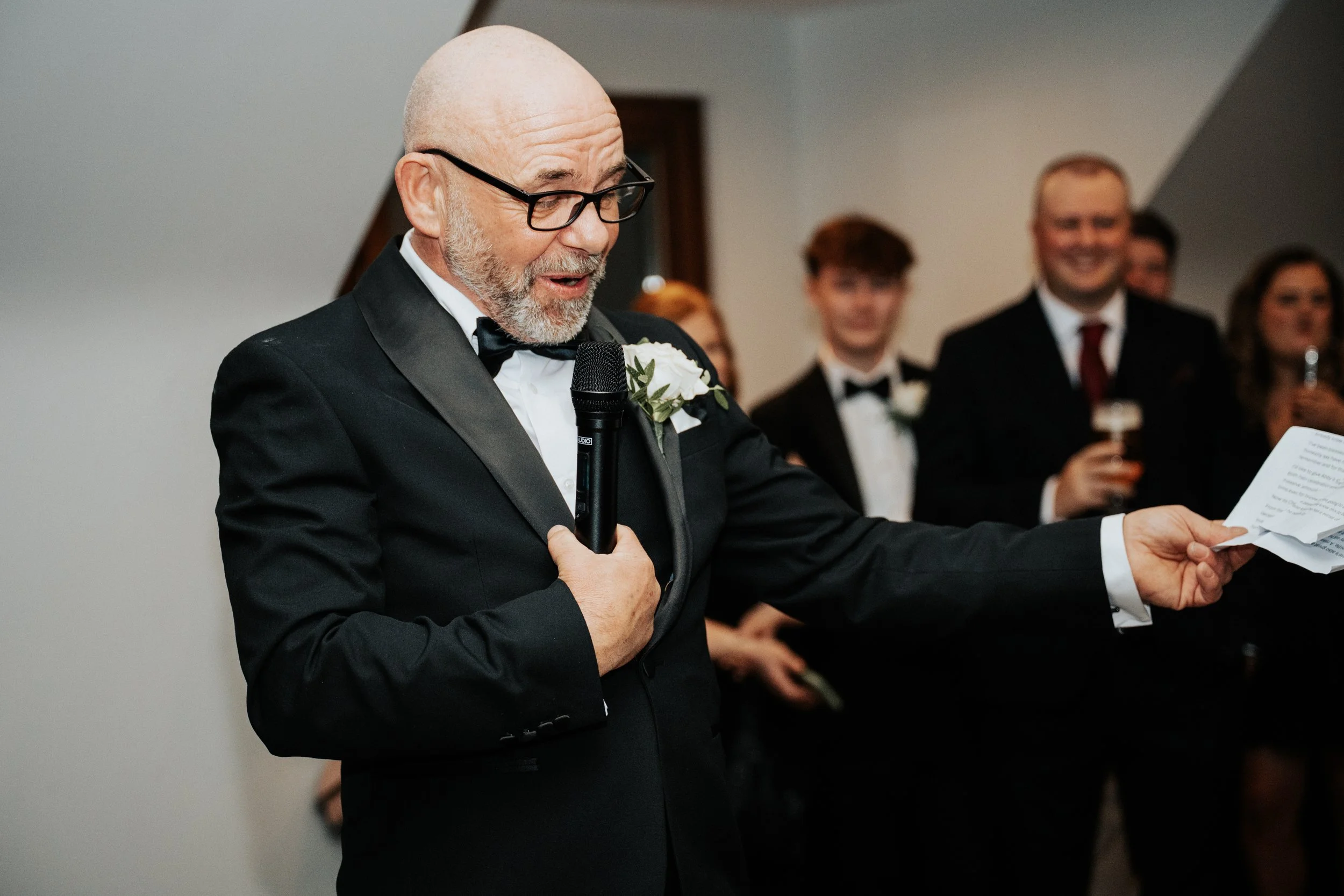 Man in tuxedo with glasses and a white boutonniere giving a speech at a wedding reception, holding a microphone in his right hand and a piece of paper in his left, with guests in formal attire in the background.