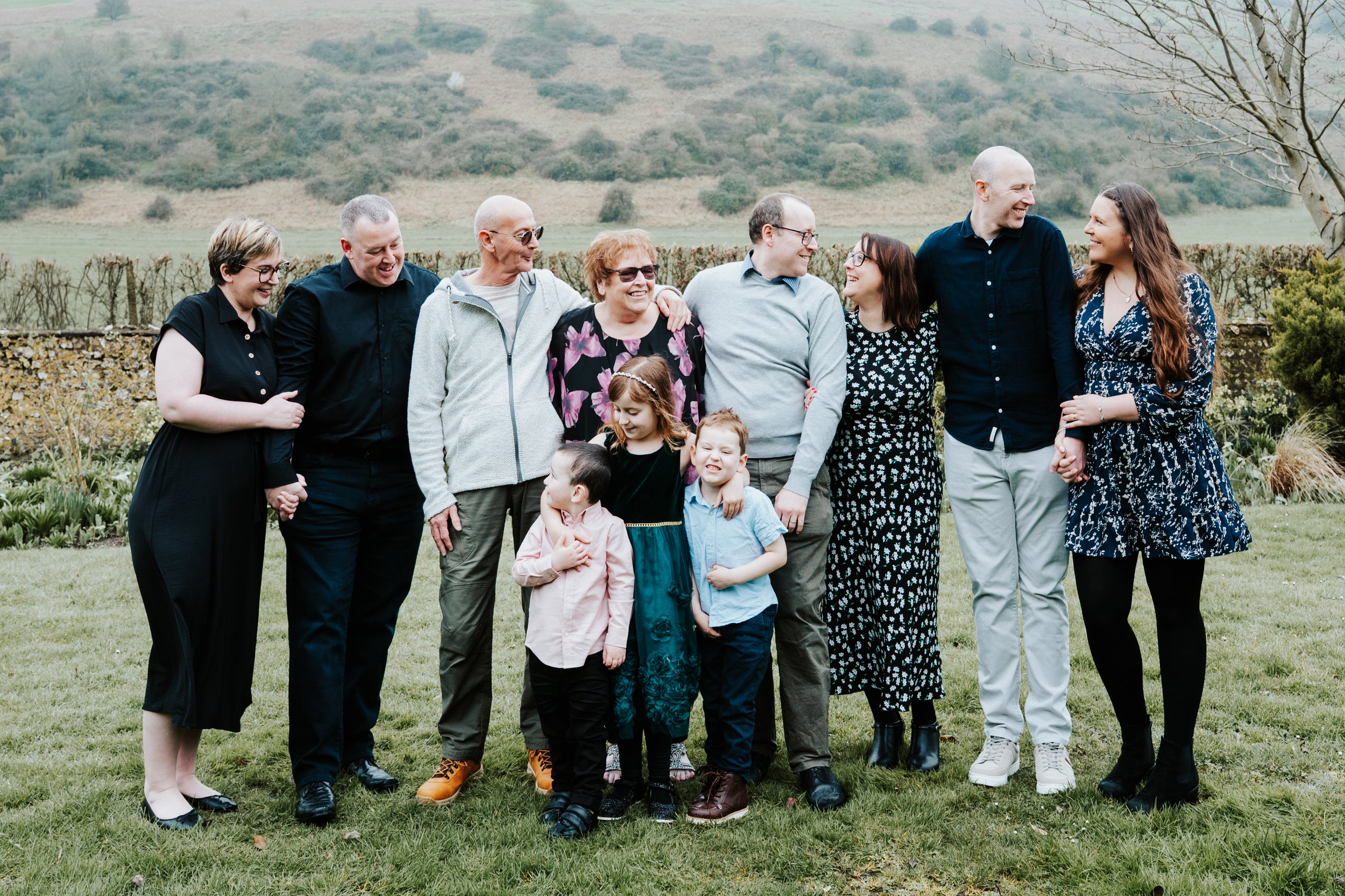 A large family standing outdoors on a grassy area with a scenic hilly background. They are smiling and holding hands.