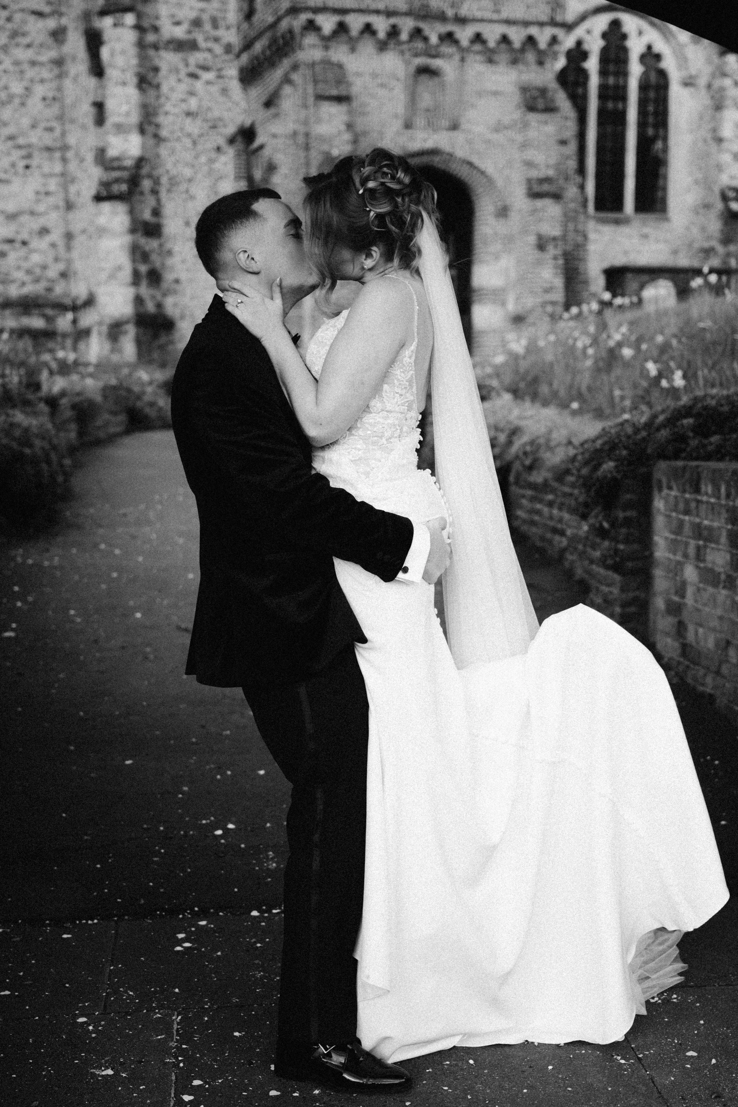 A couple sharing a kiss during their wedding, with the groom holding the bride while she wears a wedding dress and veil, in front of an old brick church.