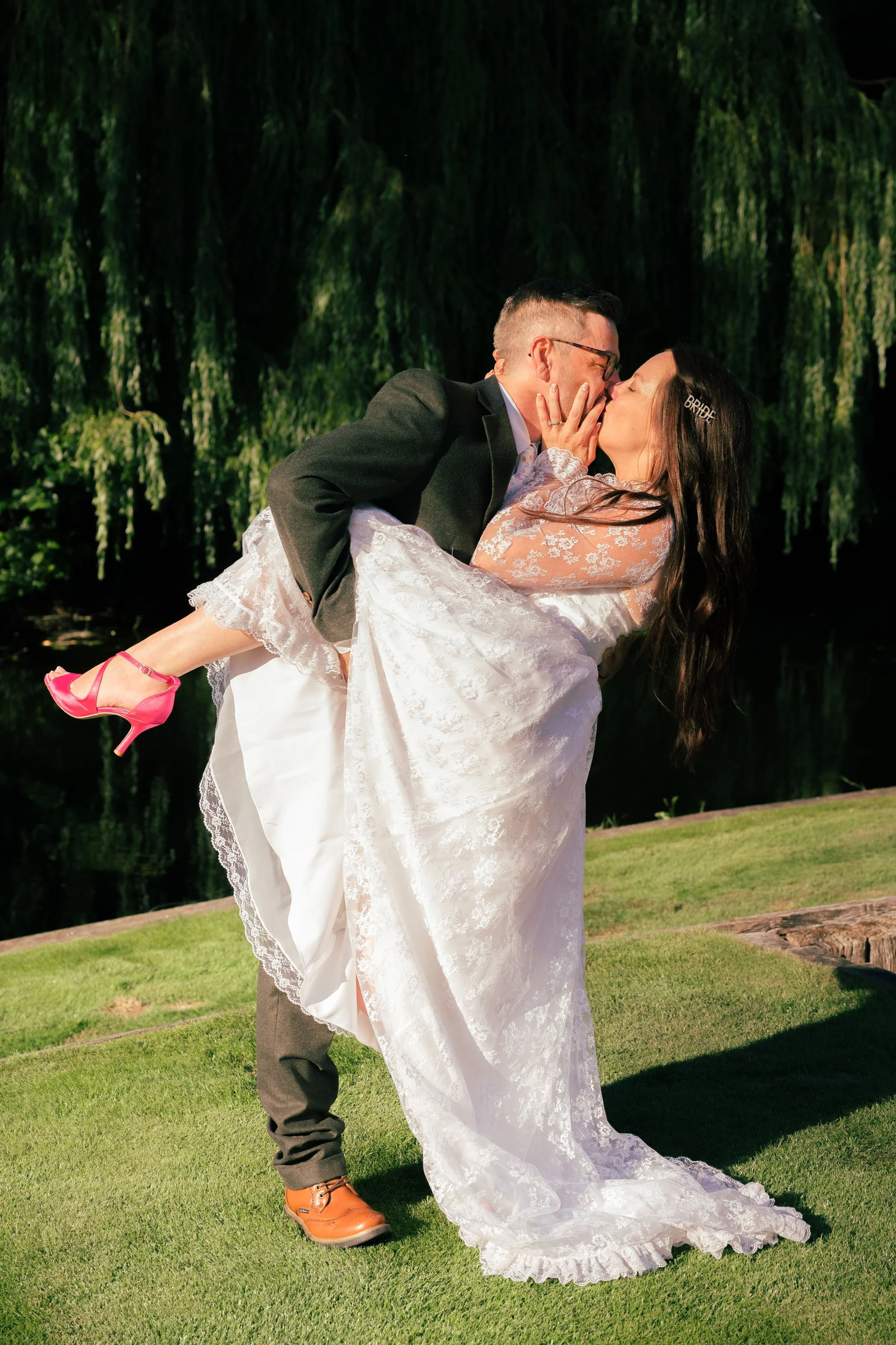 A bride and groom kissing outdoors, with the groom carrying the bride, who is wearing a white wedding gown and pink heels. The bride has a bridal hair clip that says "BRIDE" and is dressed in a lace gown, while the groom is in a dark jacket and brown