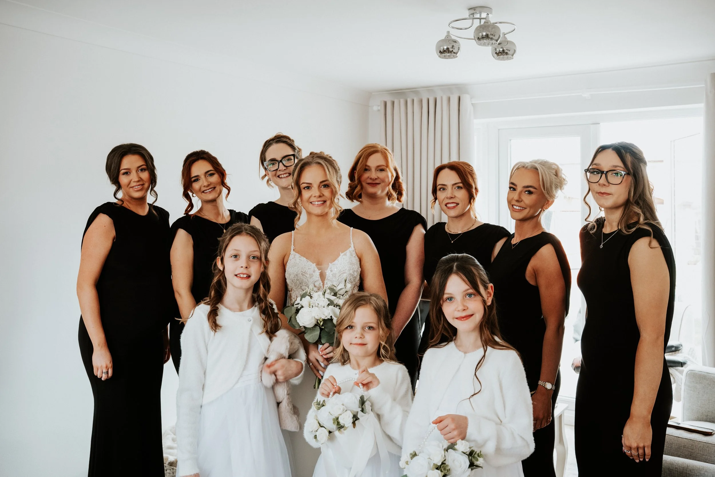 Group of women and young girls dressed for a wedding in a bright room with white walls, some in black dresses and three in white dresses holding bouquets, smiling for a photo.