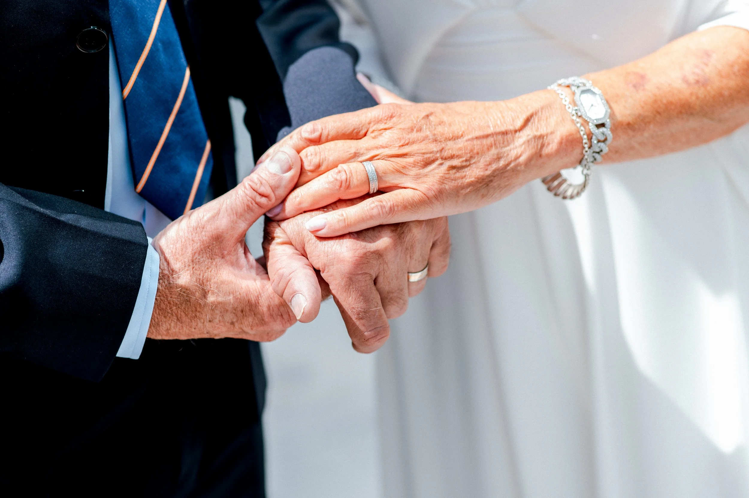 An elderly man and woman holding hands, with the woman's hand resting on top of the man's hand, both wearing wedding rings, in a close-up shot.