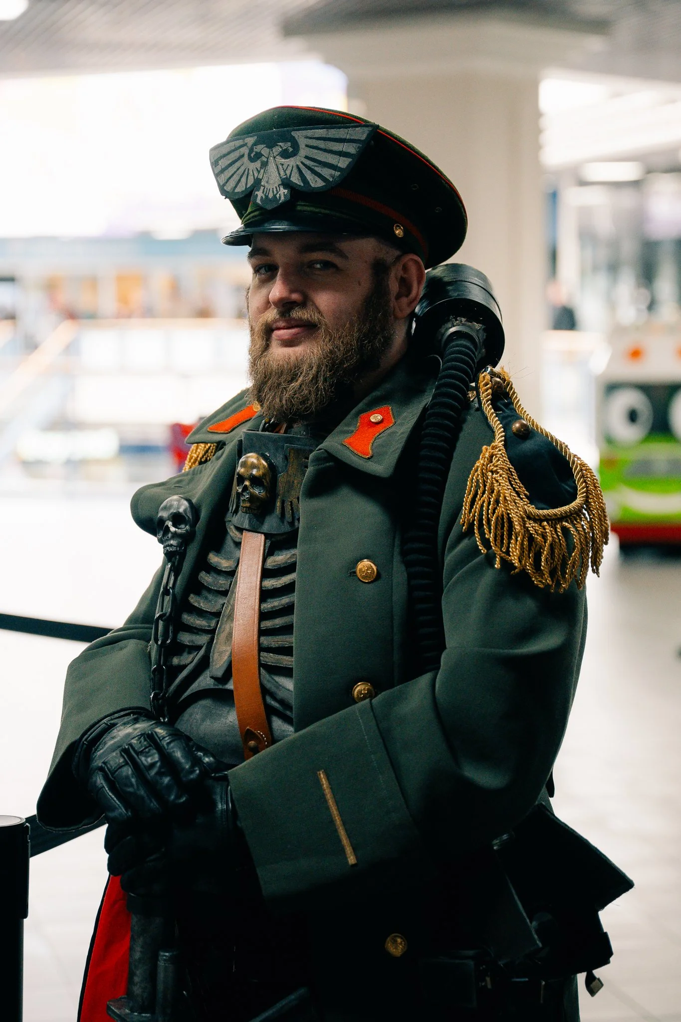 A man dressed in a detailed medieval or fantasy soldier costume, including a hat with an eagle and skull embellishments, a green tunic with gold accents, and gloves, standing indoors with a bus in the background.