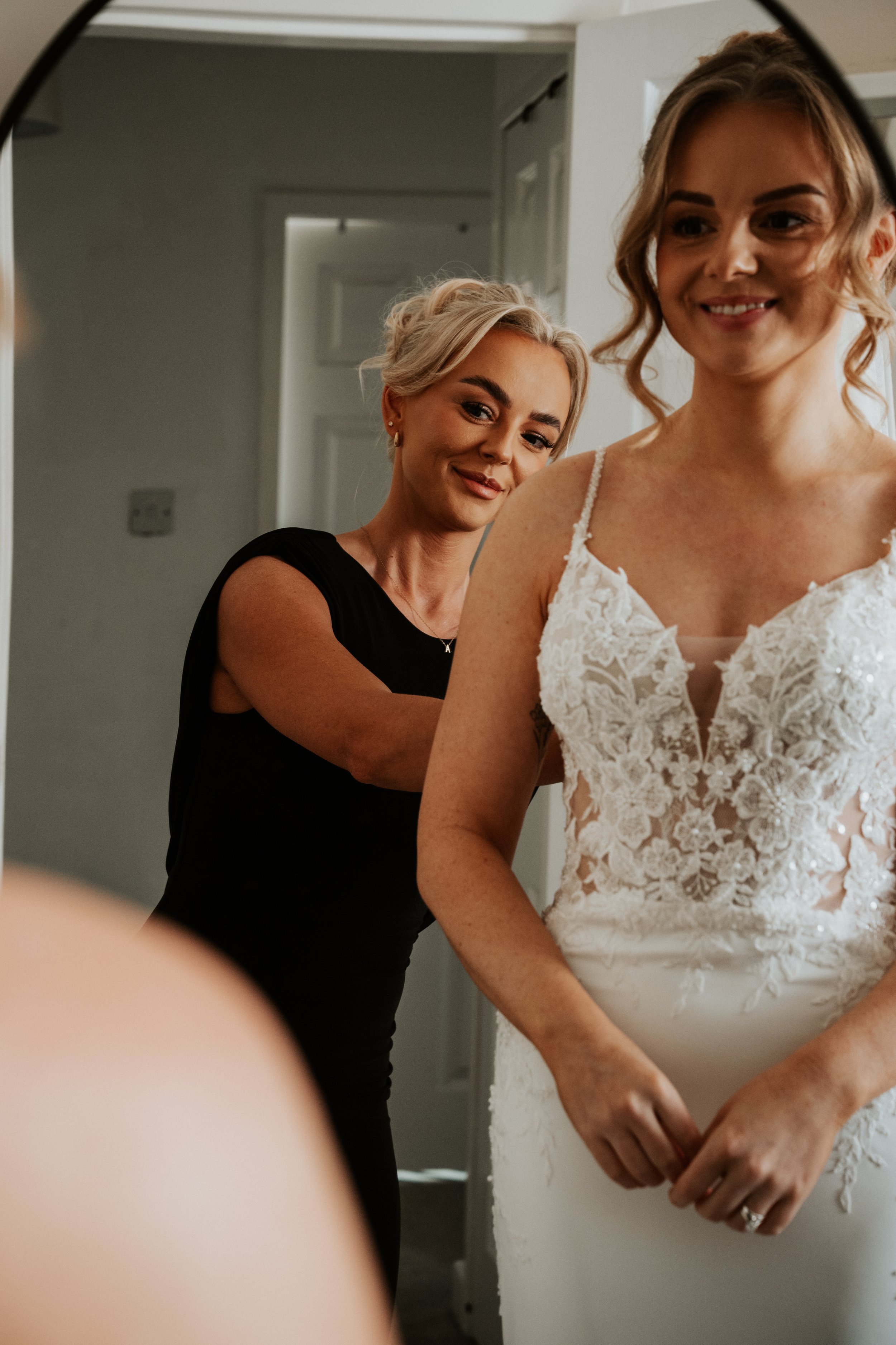 A woman helping a bride get ready, with the bride smiling in her wedding dress, visible lace detailing, and wedding ring.