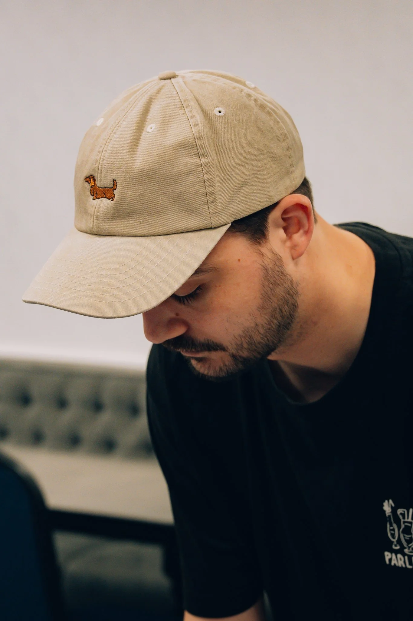 A man wearing a beige baseball cap with a small embroidered dachshund dog on the front. He is looking downward and has a beard, wearing a black T-shirt, sitting indoors with a gray cushioned bench in the background.