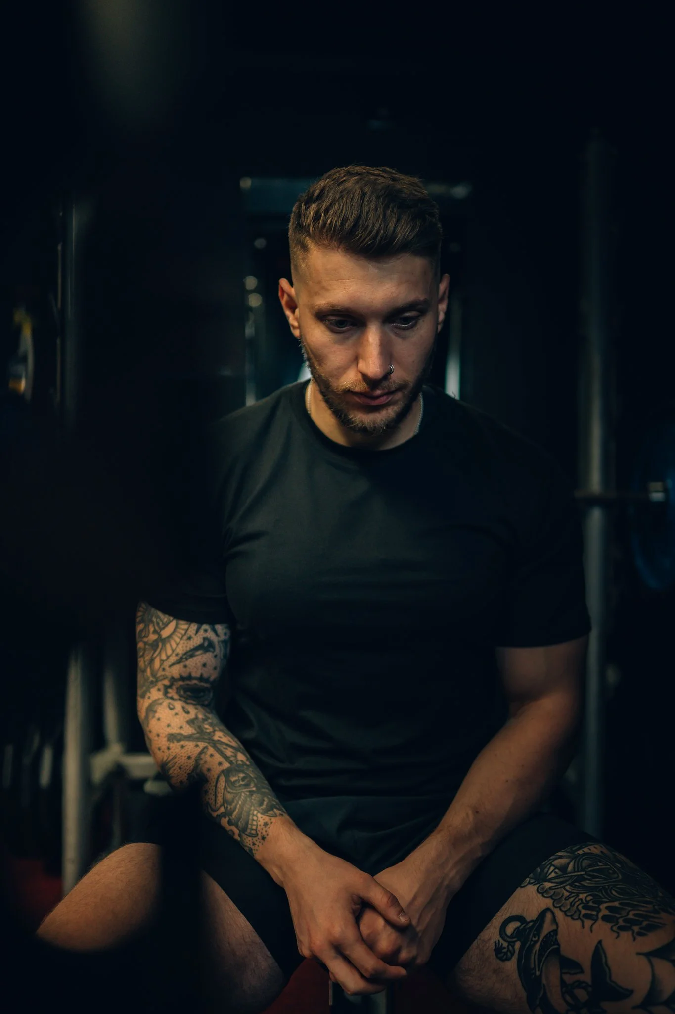 A young man with tattoos and a nose ring sitting in a gym, looking down with a focused expression.