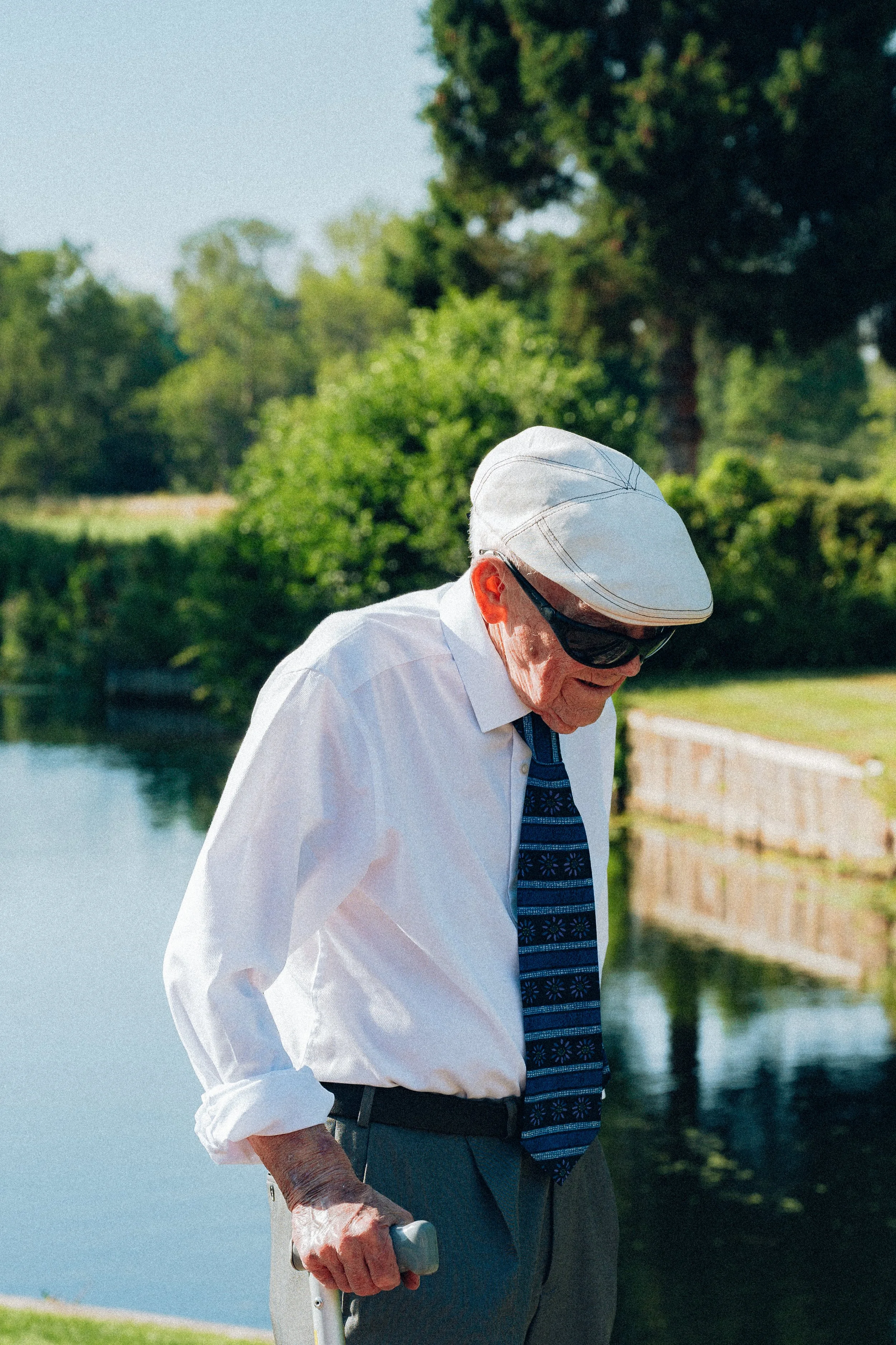 An elderly man wearing a white cap, sunglasses, a white button-down shirt, a patterned tie, and gray pants, stands near a body of water holding a cane.