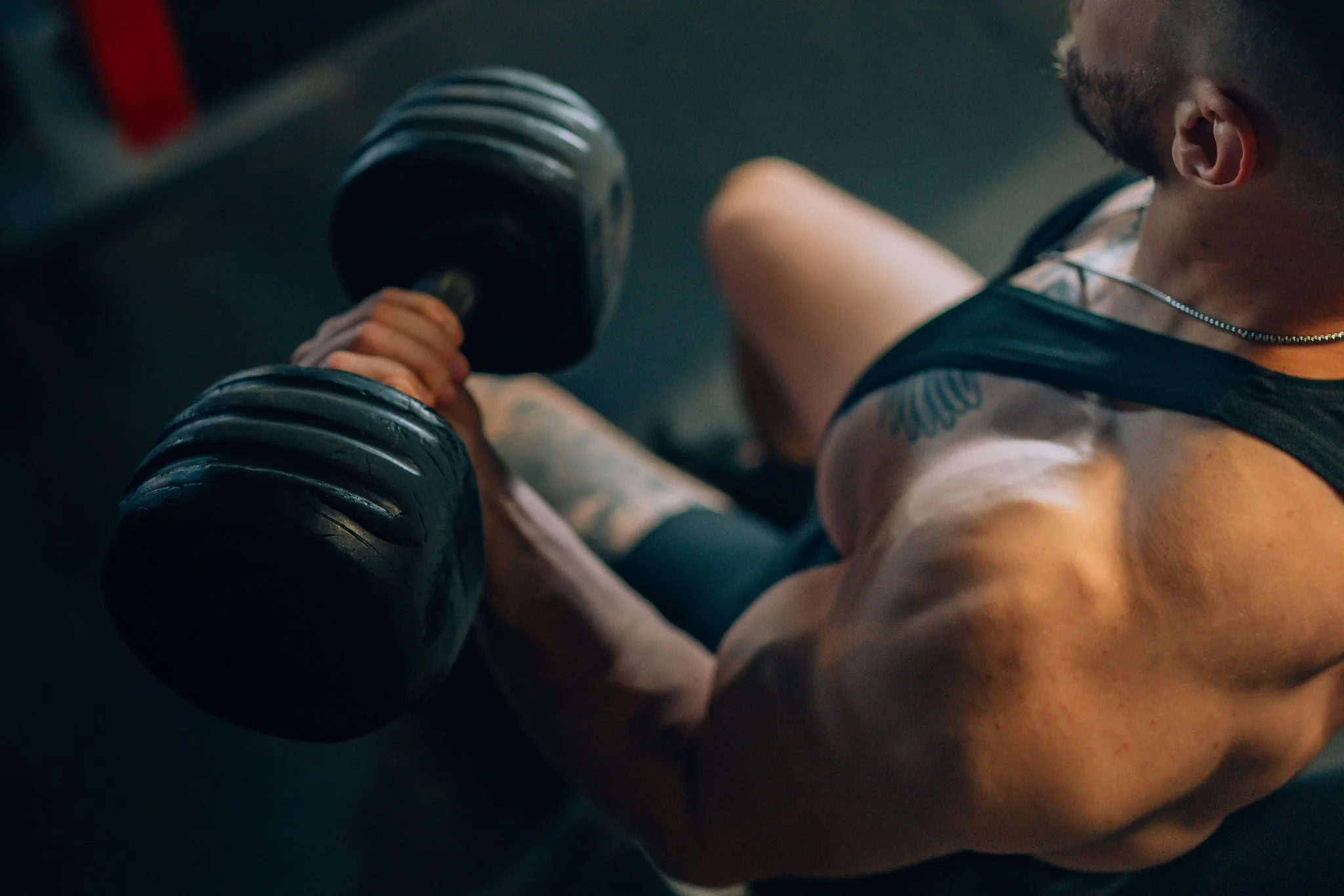 A muscular man in a gym, wearing a tank top, is performing an exercise with a large dumbbell in a seated position.