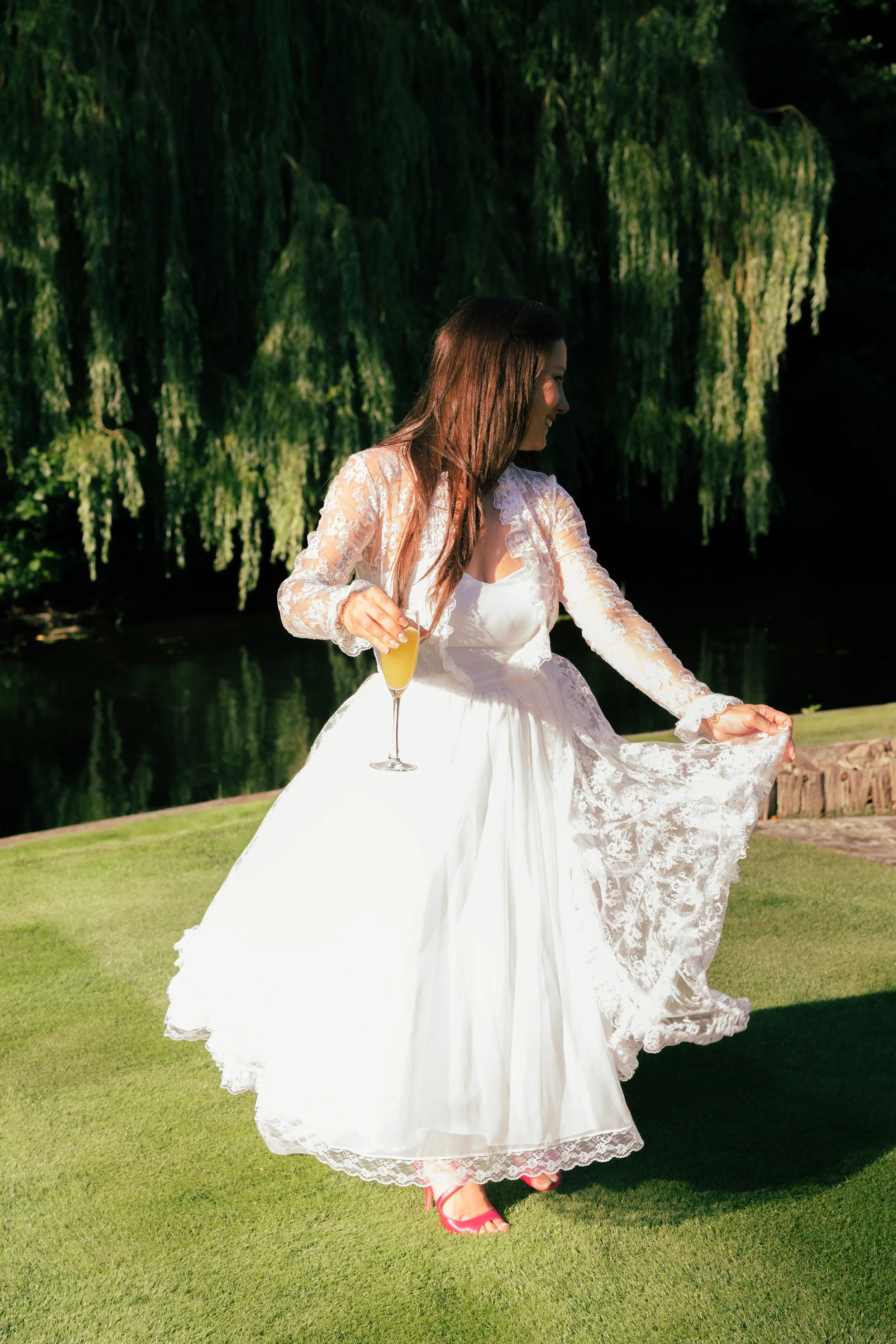Woman in white wedding dress holding a glass of champagne outdoors, standing on grass near a body of water with hanging green trees in the background.