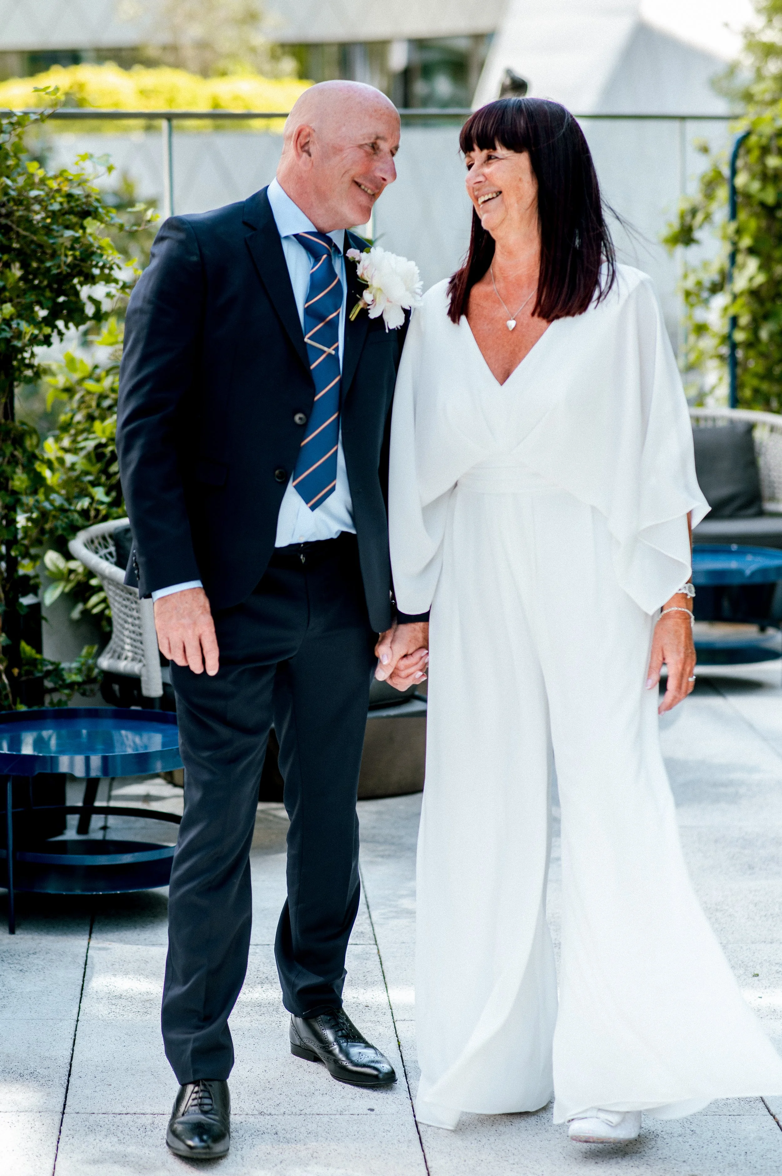 A man and woman, dressed in formal attire, holding hands and smiling at each other outdoors in a garden setting.