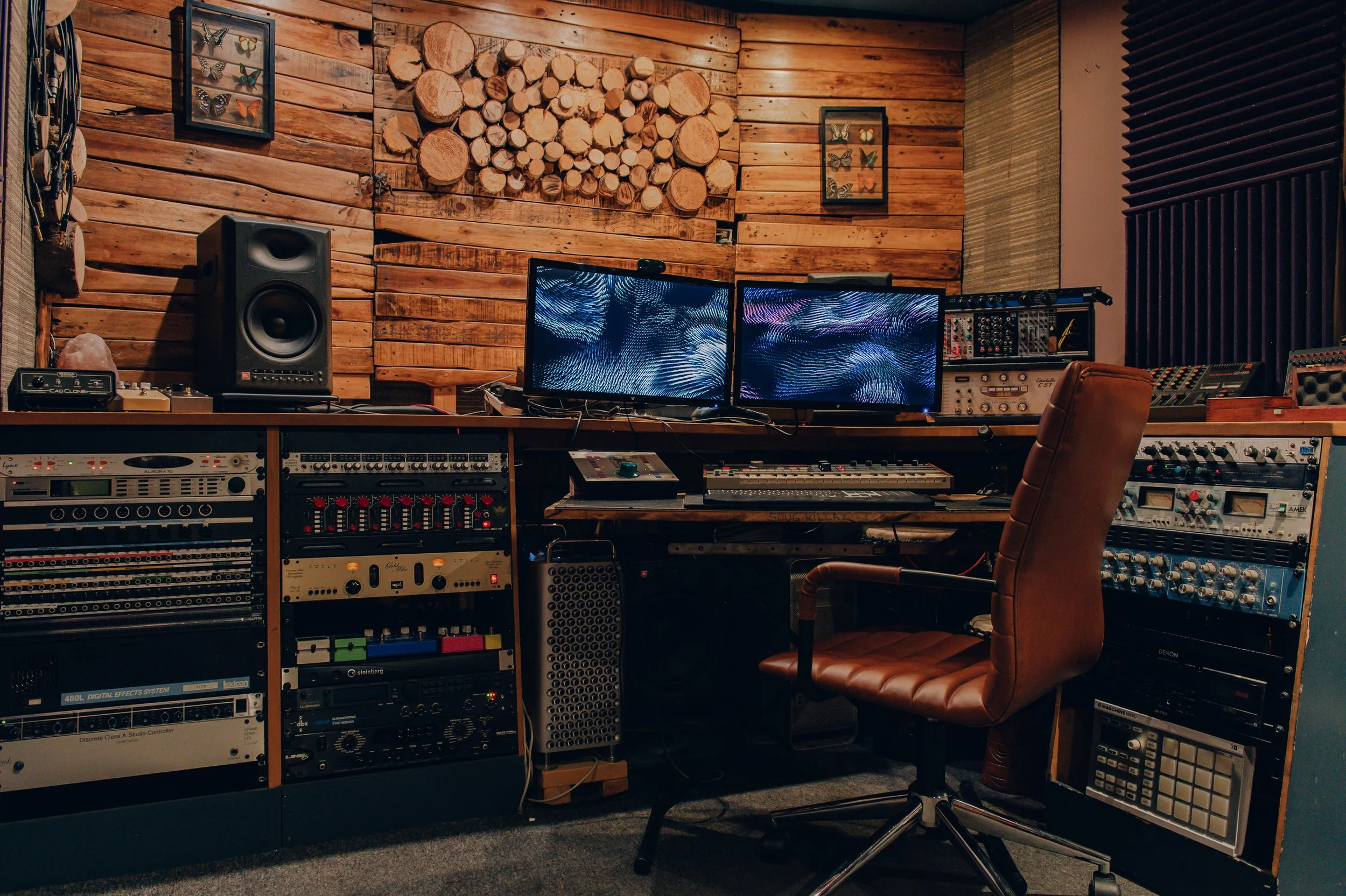 Music recording studio with wooden wall and two computer monitors displaying abstract visuals. Equipment racks with audio gear and a brown leather swivel chair.