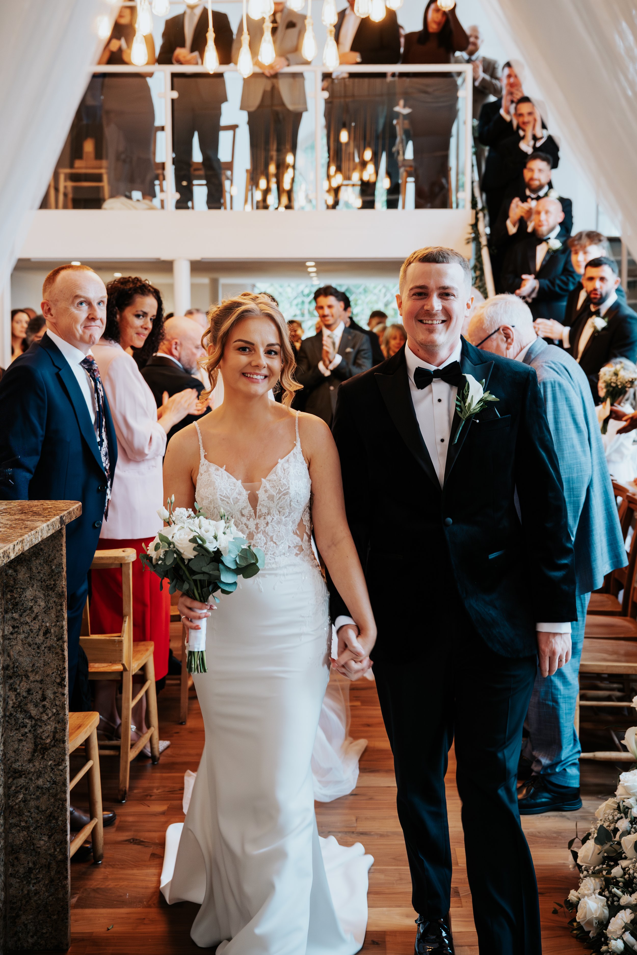 A newly married couple walking down the aisle at their wedding ceremony, holding hands, with guests clapping and smiling around them.
