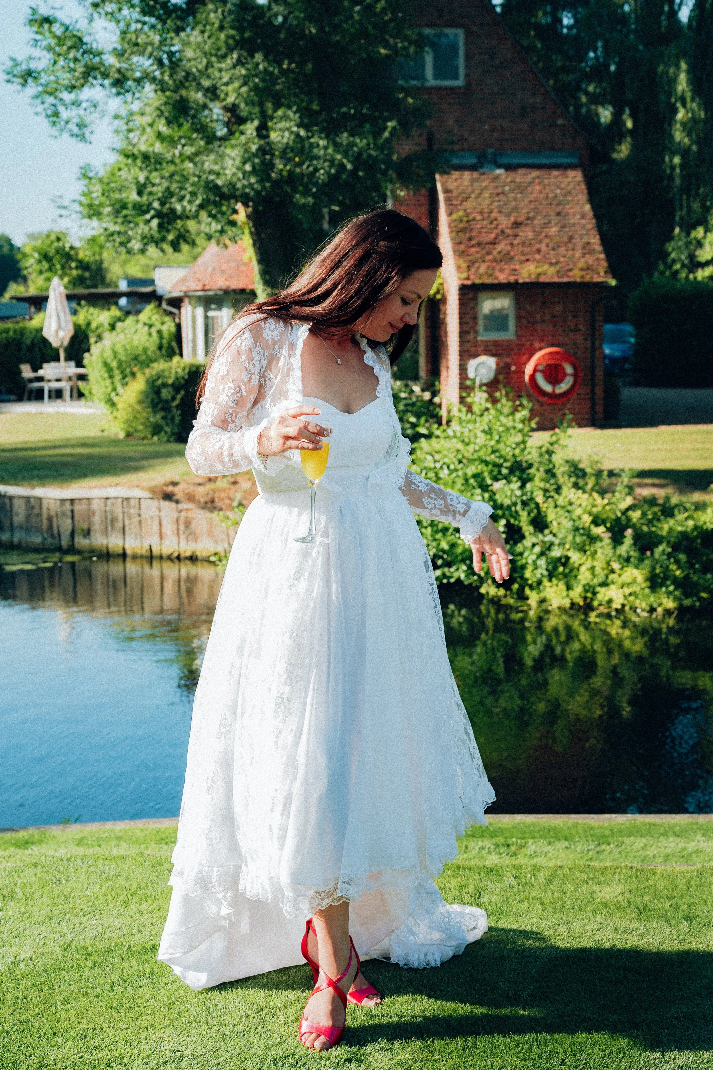 A woman in a white bridal dress with lace sleeves standing on a green lawn near a pond, holding a glass of champagne, wearing red high heels, with trees and a brick building in the background on a sunny day.