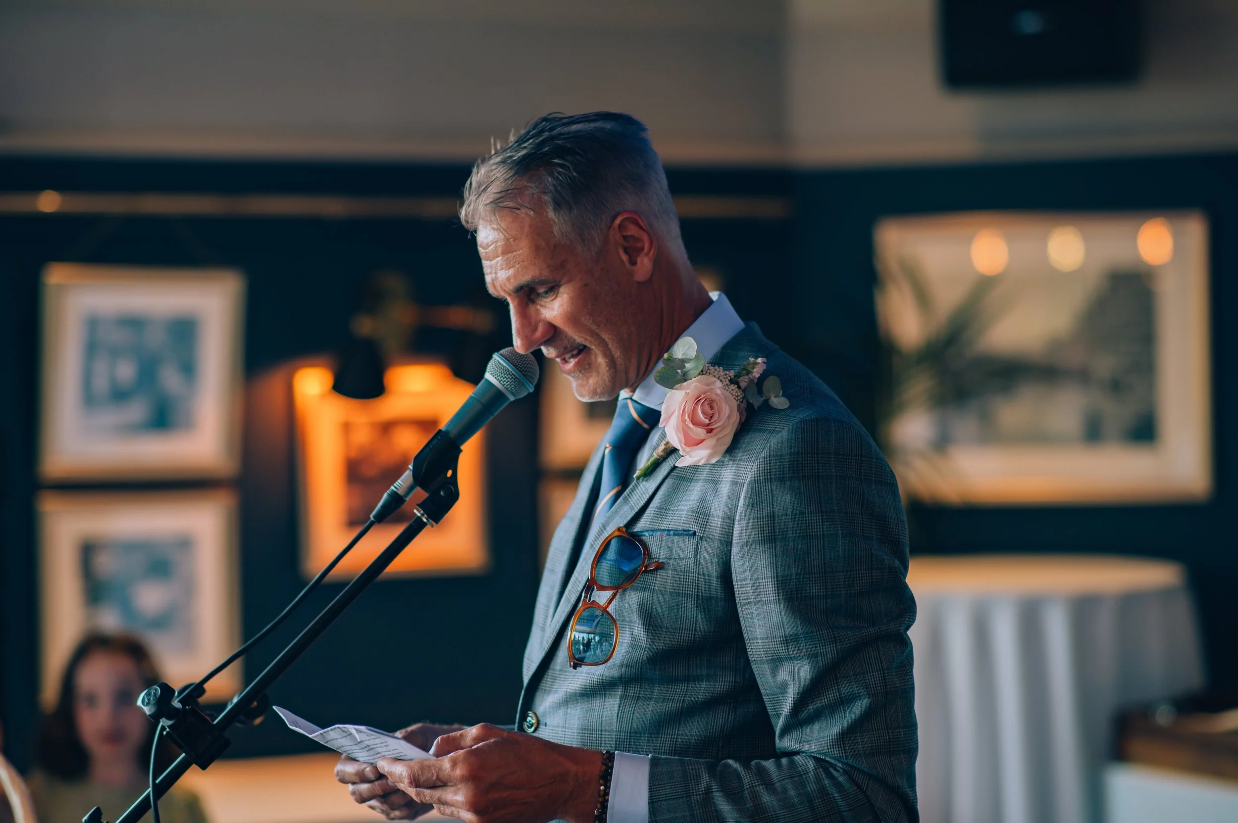 A man in a gray plaid suit giving a speech at a wedding reception, with a pink rose boutonniere on his lapel and reading from a piece of paper, in a warmly lit room decorated with framed photos and artwork.