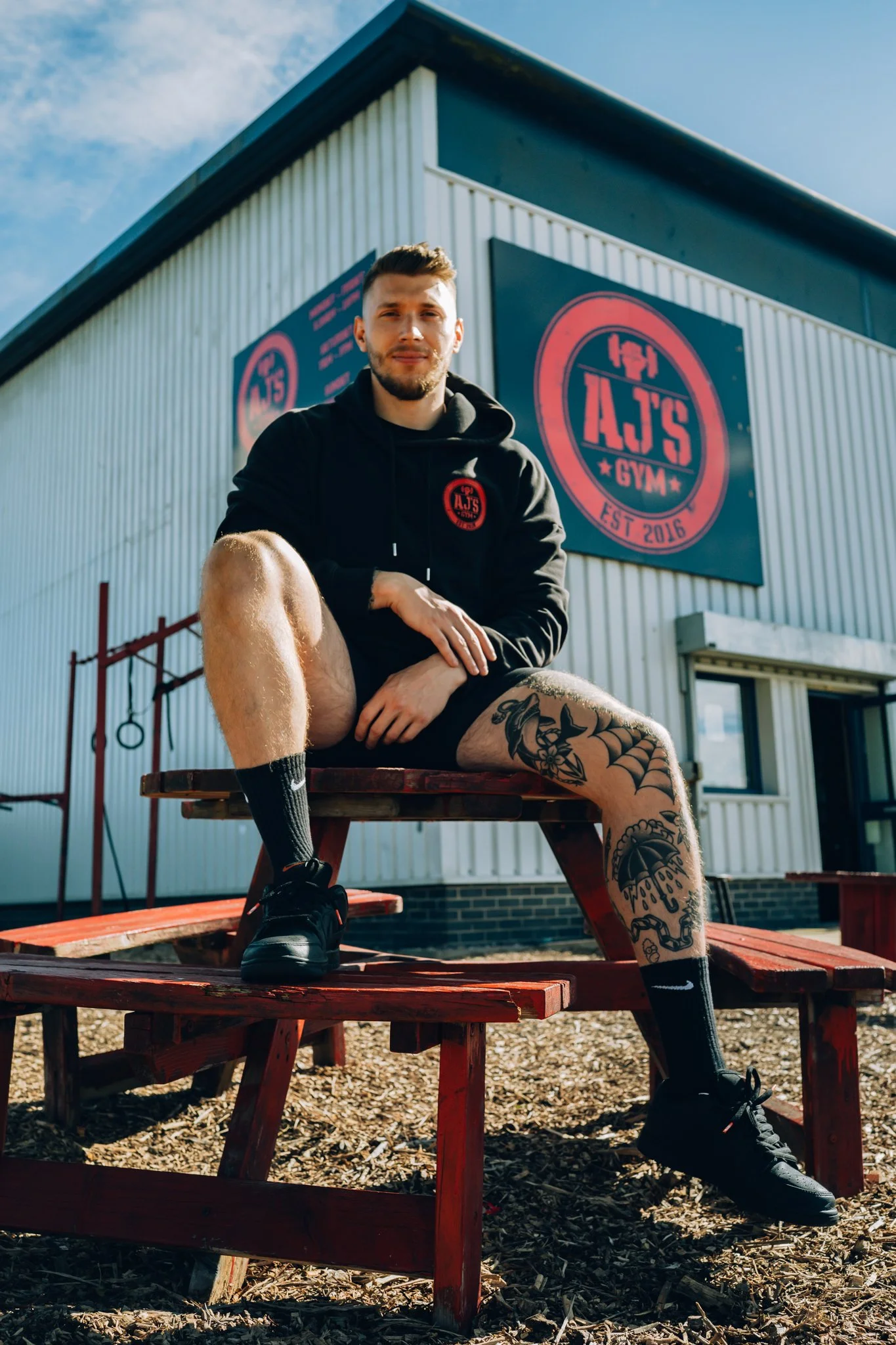 A young man with tattoos, wearing a black hoodie, black shorts, black socks, and athletic shoes, sitting on a wooden picnic table outside a gym named AJ's Gym, with gym equipment and a large sign visible on the building.