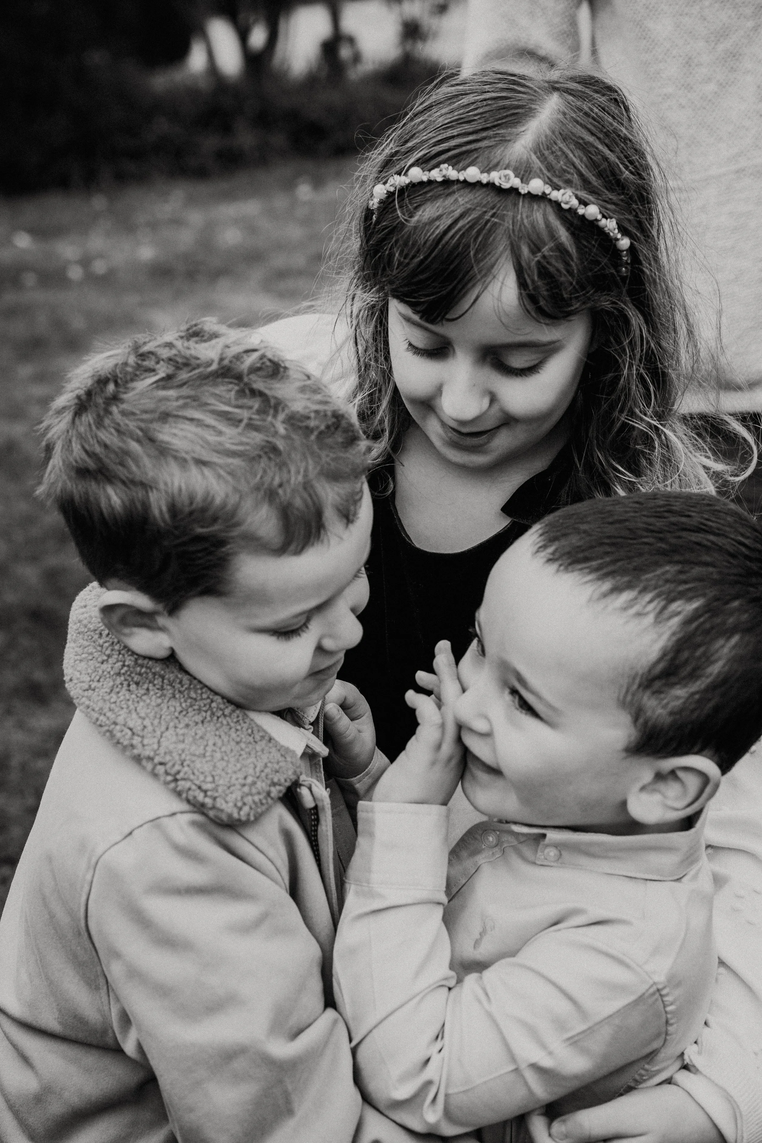 A black-and-white photo of a young girl with curly hair and a headband, looking down at two young boys who are touching their faces and smiling at each other.