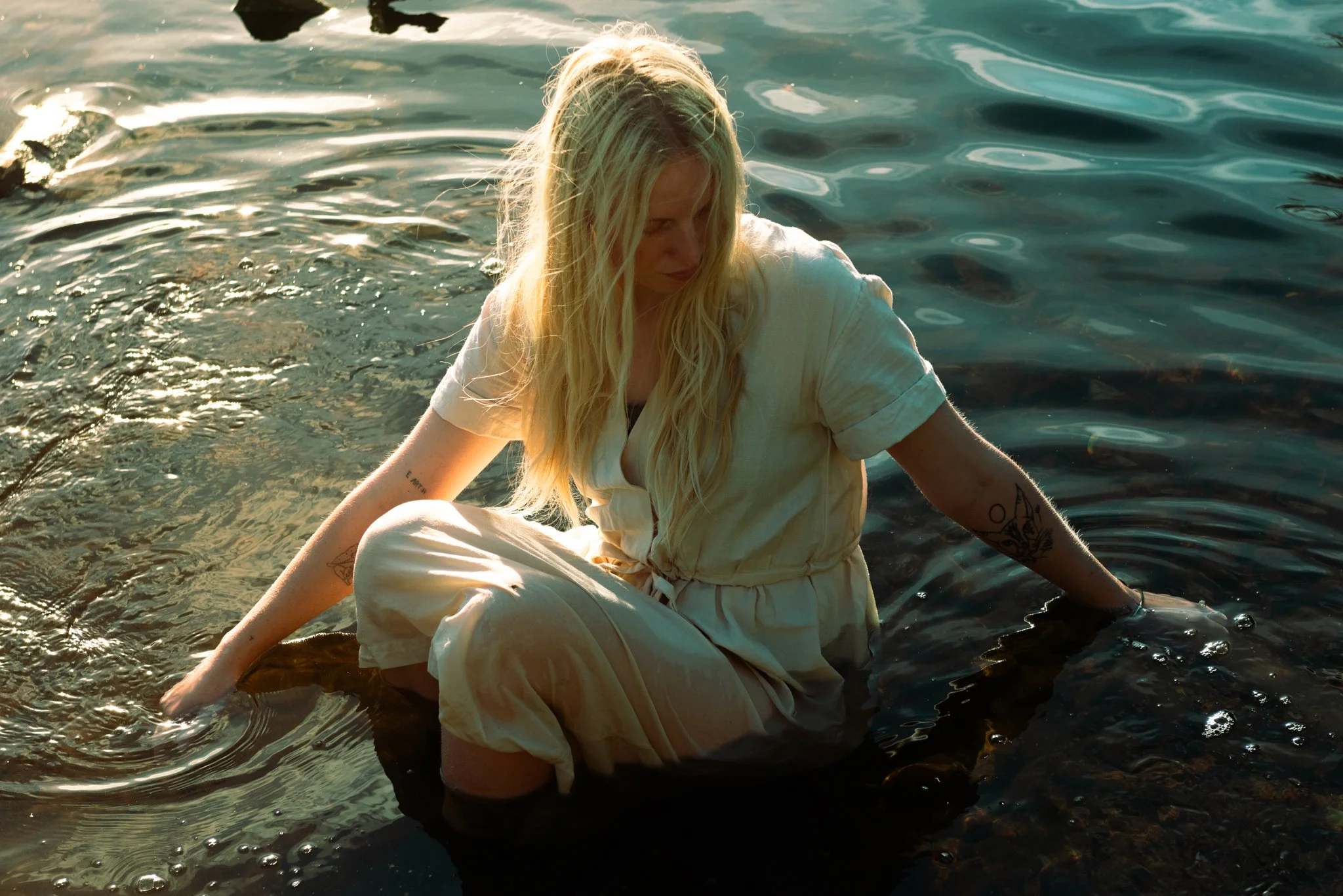 A blonde woman in a light-colored dress sitting in shallow water with her legs crossed, holding onto a rock, and looking down, with the water reflecting the sunlight.
