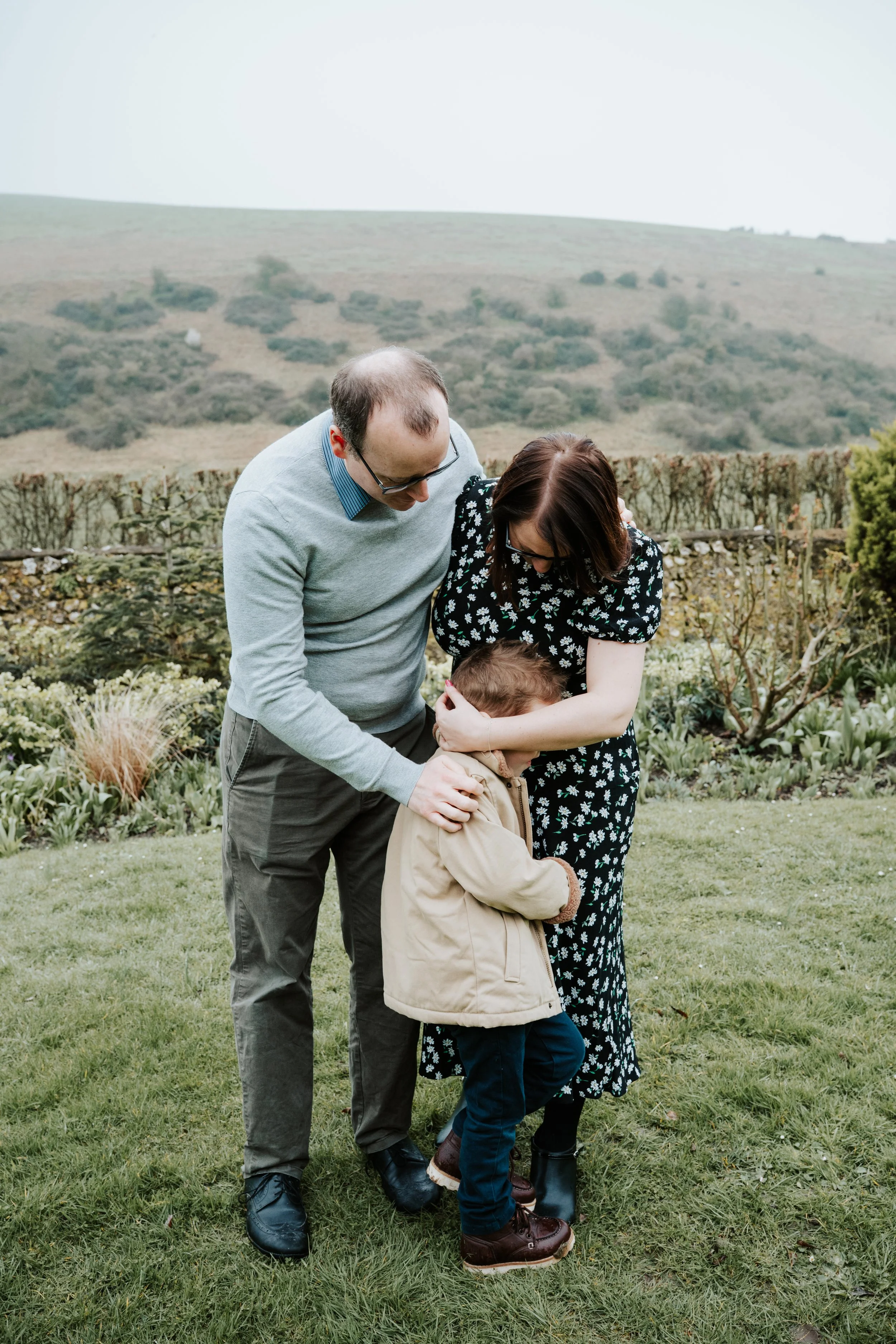 A family of three standing on a grassy lawn outdoors, with a view of rolling hills in the background. The man is leaning over a young boy, who appears to be upset, and the woman is comforting the boy by holding his face and hugging him. The boy is we