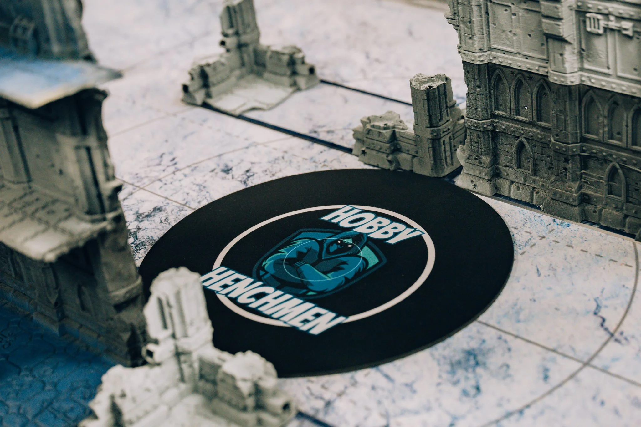 Tabletop gaming terrain with miniature buildings and a circular black game mat with a logo featuring a ninja and the text "Hobby Fencing".
