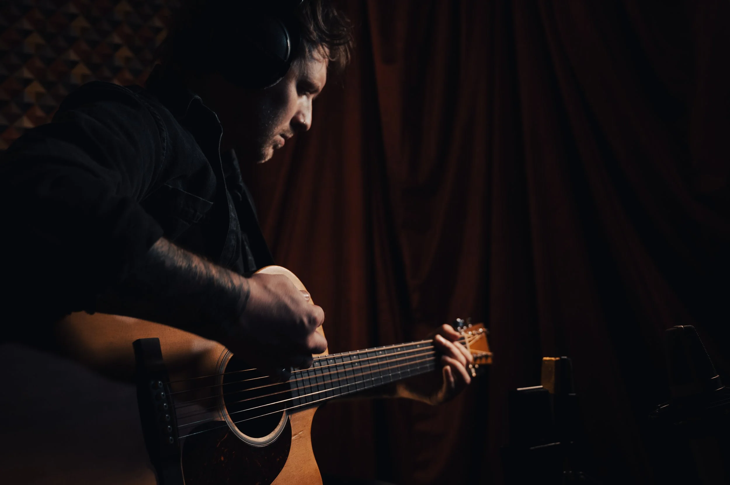 A man wearing headphones playing an acoustic guitar in a dimly lit recording studio.