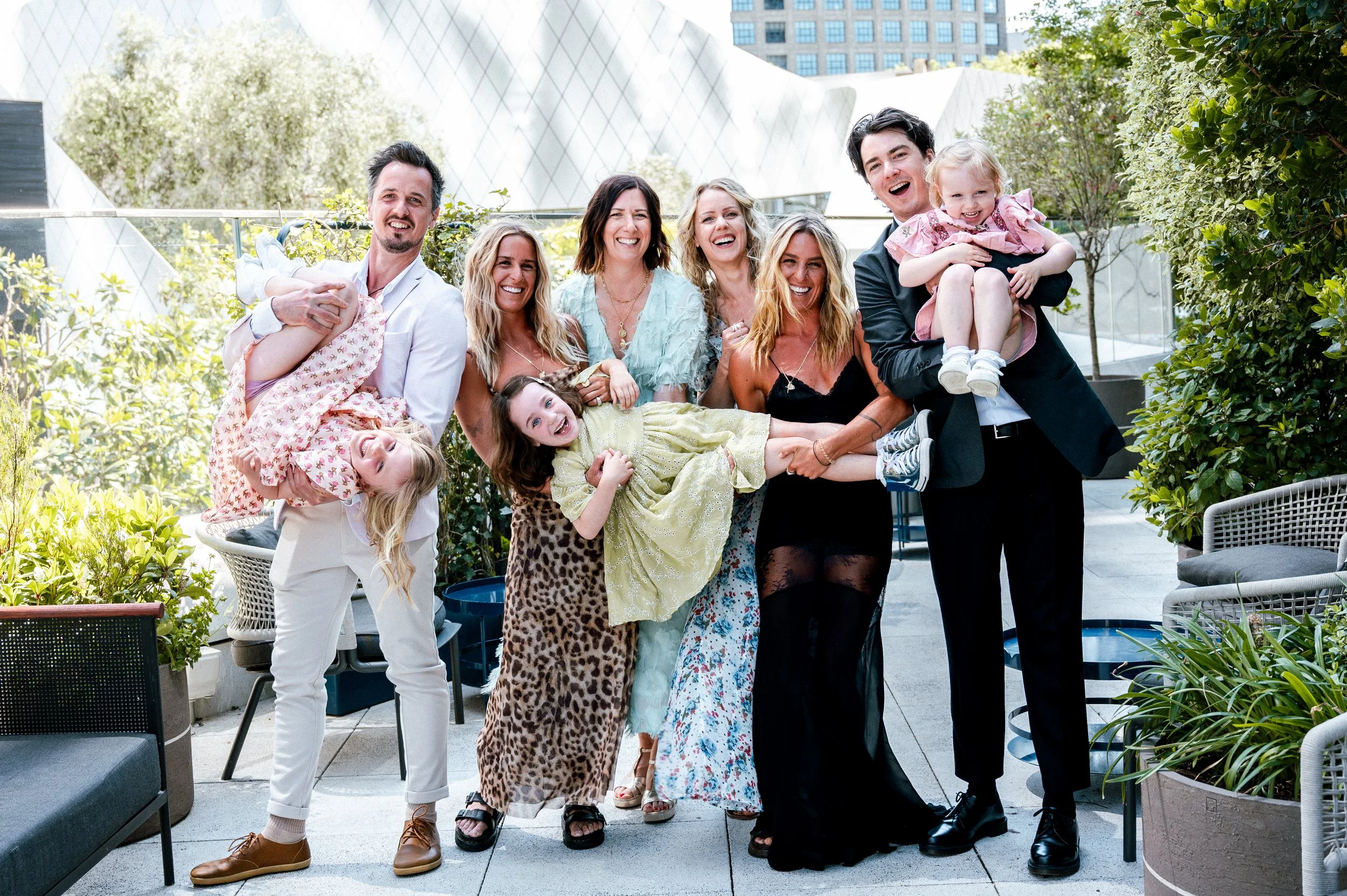 A group of nine people, including children, enjoying a sunny outdoor gathering in a garden or patio area, smiling and holding each other.
