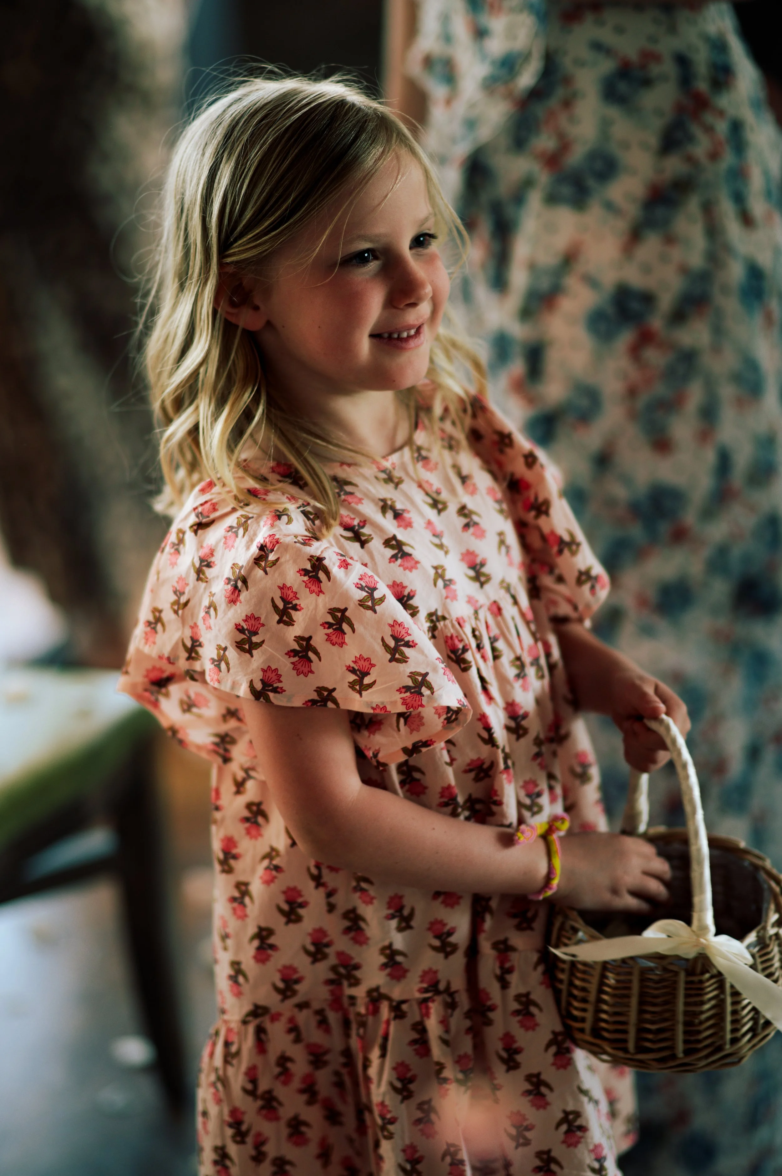 A young girl with blonde hair smiling, wearing a pink dress with a floral pattern, holding a small basket with a ribbon.