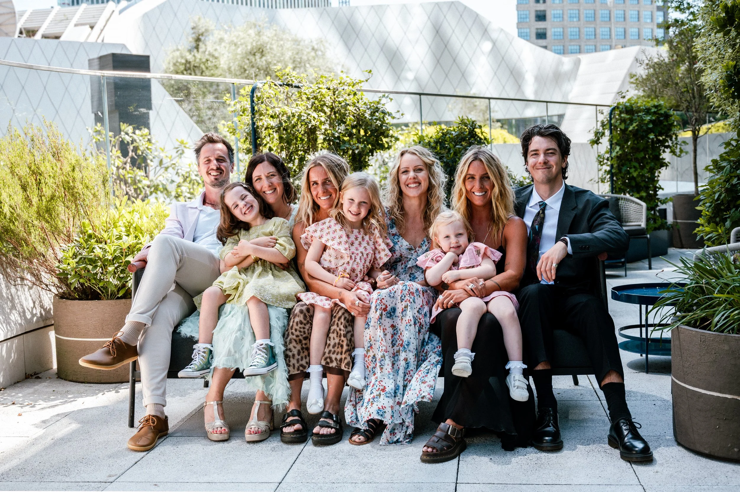 A group of adults and children sitting together on a bench outdoors, smiling. The setting is a sunny urban rooftop with plants and modern buildings in the background.