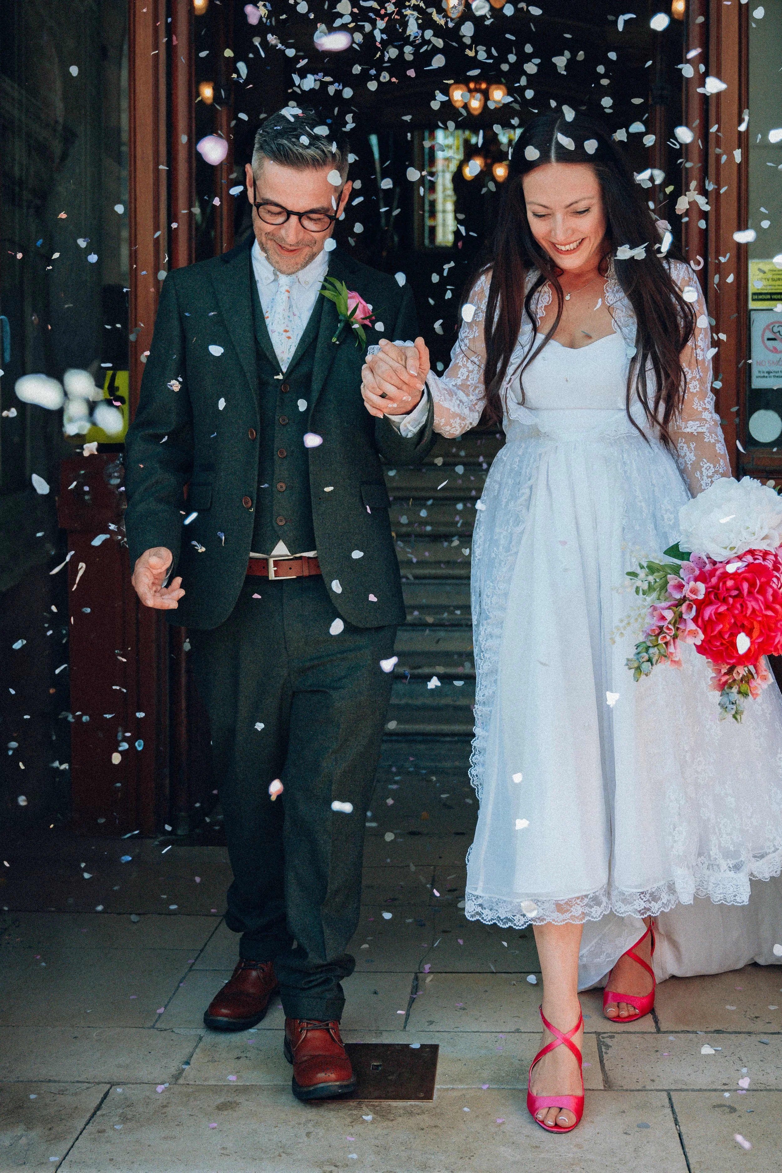 A newlywed couple walking out of a building, holding hands and smiling, surrounded by falling confetti during their wedding celebration.