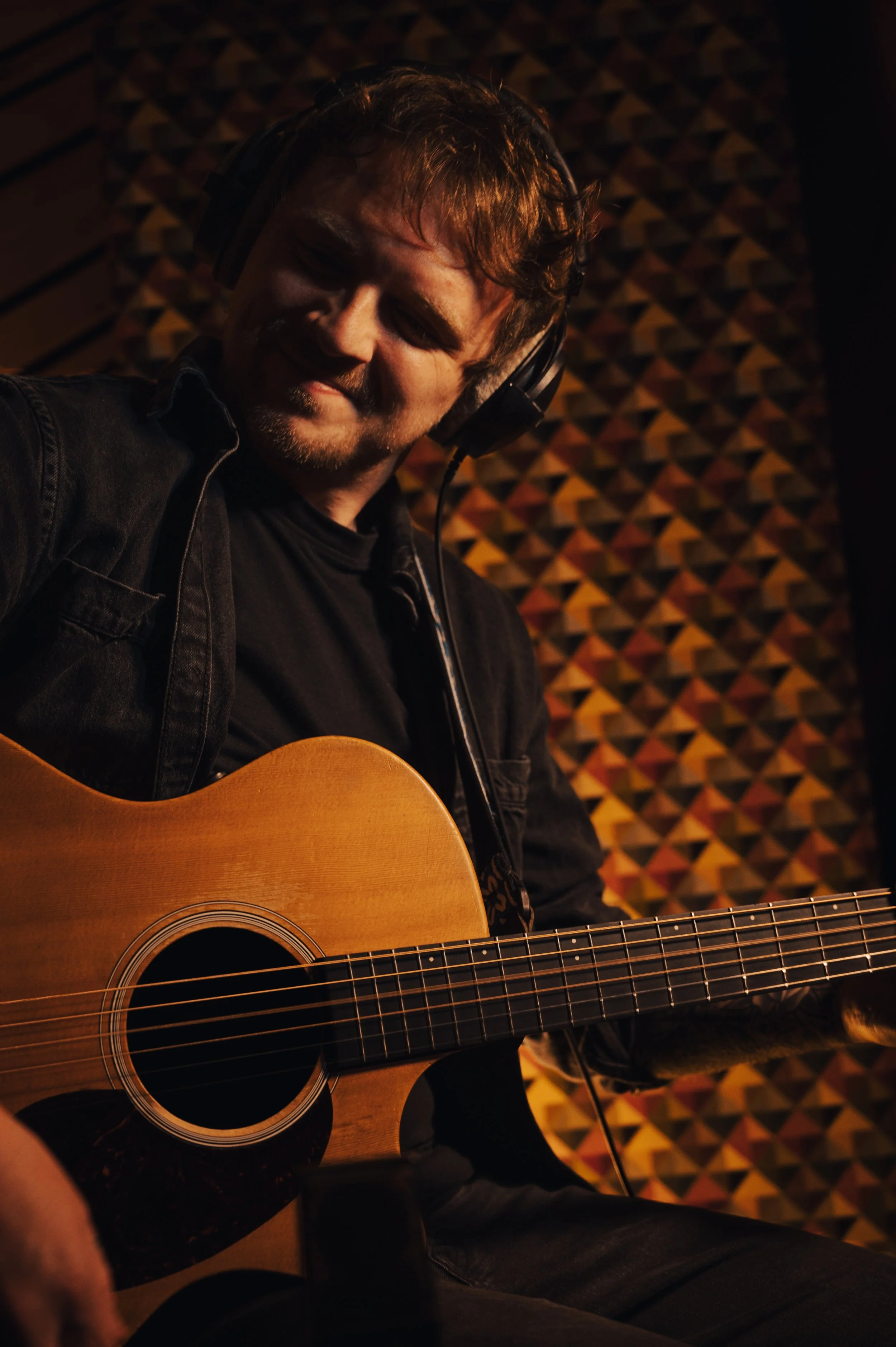 A man with headphones playing an acoustic guitar in a recording studio.