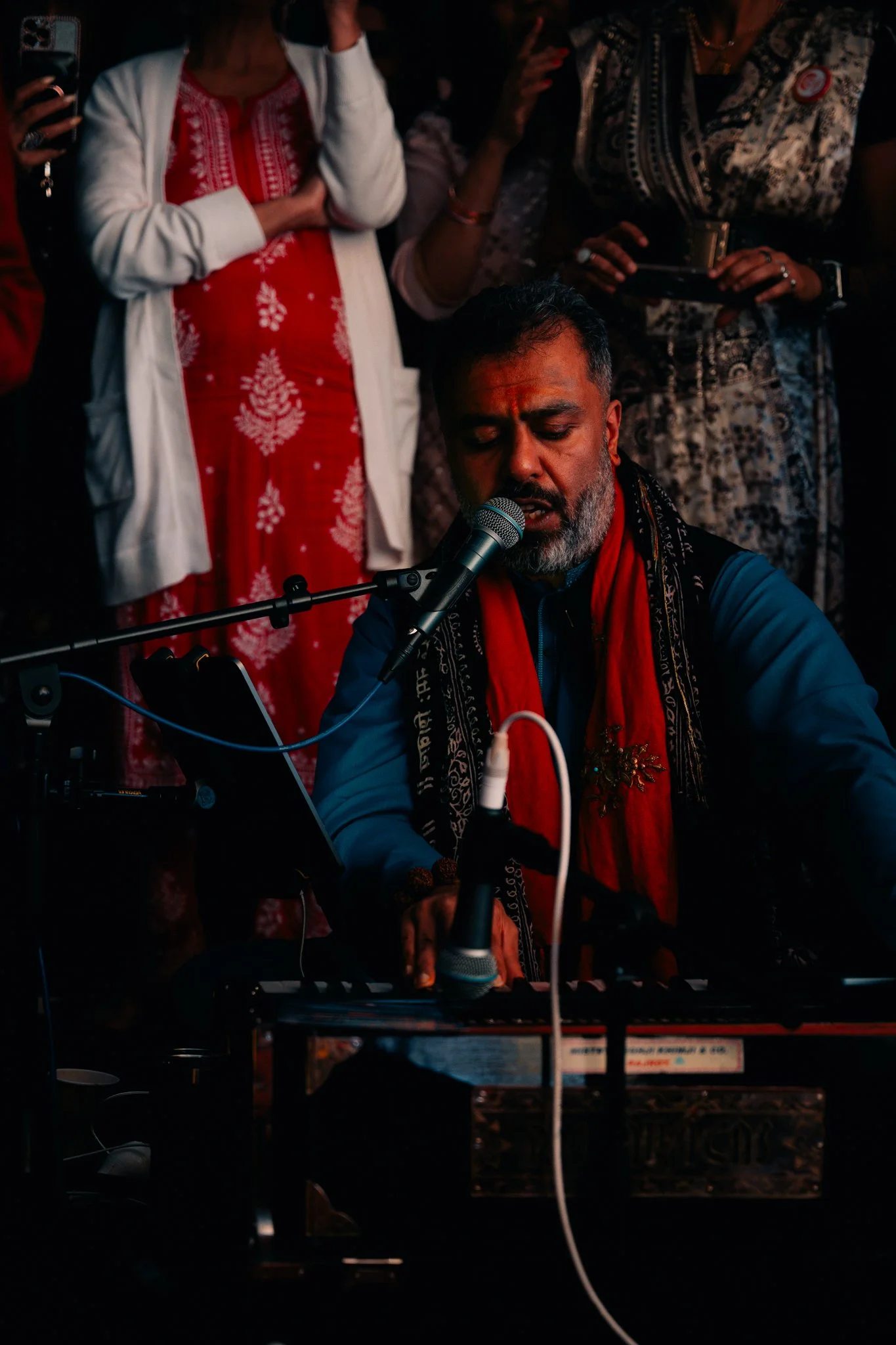 A man with dark hair and a beard, wearing a blue shirt and red scarf, singing into a microphone while playing a keyboard. Several people are standing behind him, watching, with some holding phones.