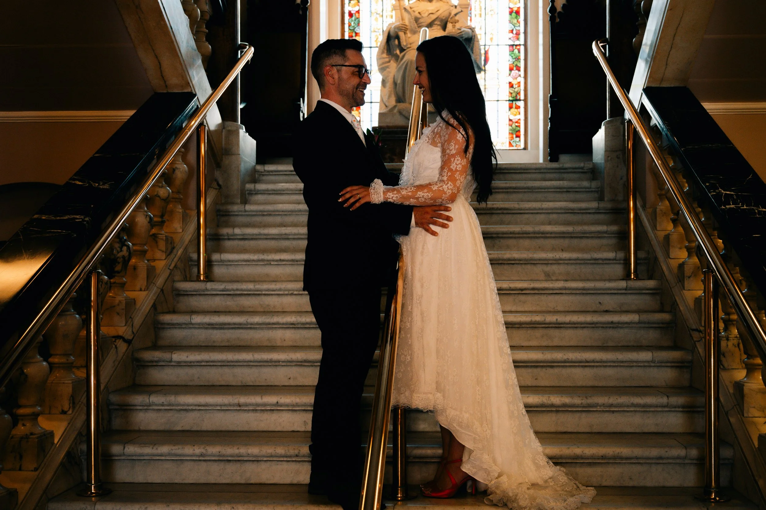 A wedding couple standing on a marble staircase inside a building with a stained glass window in the background. The bride is wearing a long white lace wedding dress with lace sleeves and red heels. The groom is wearing a black suit and glasses. They