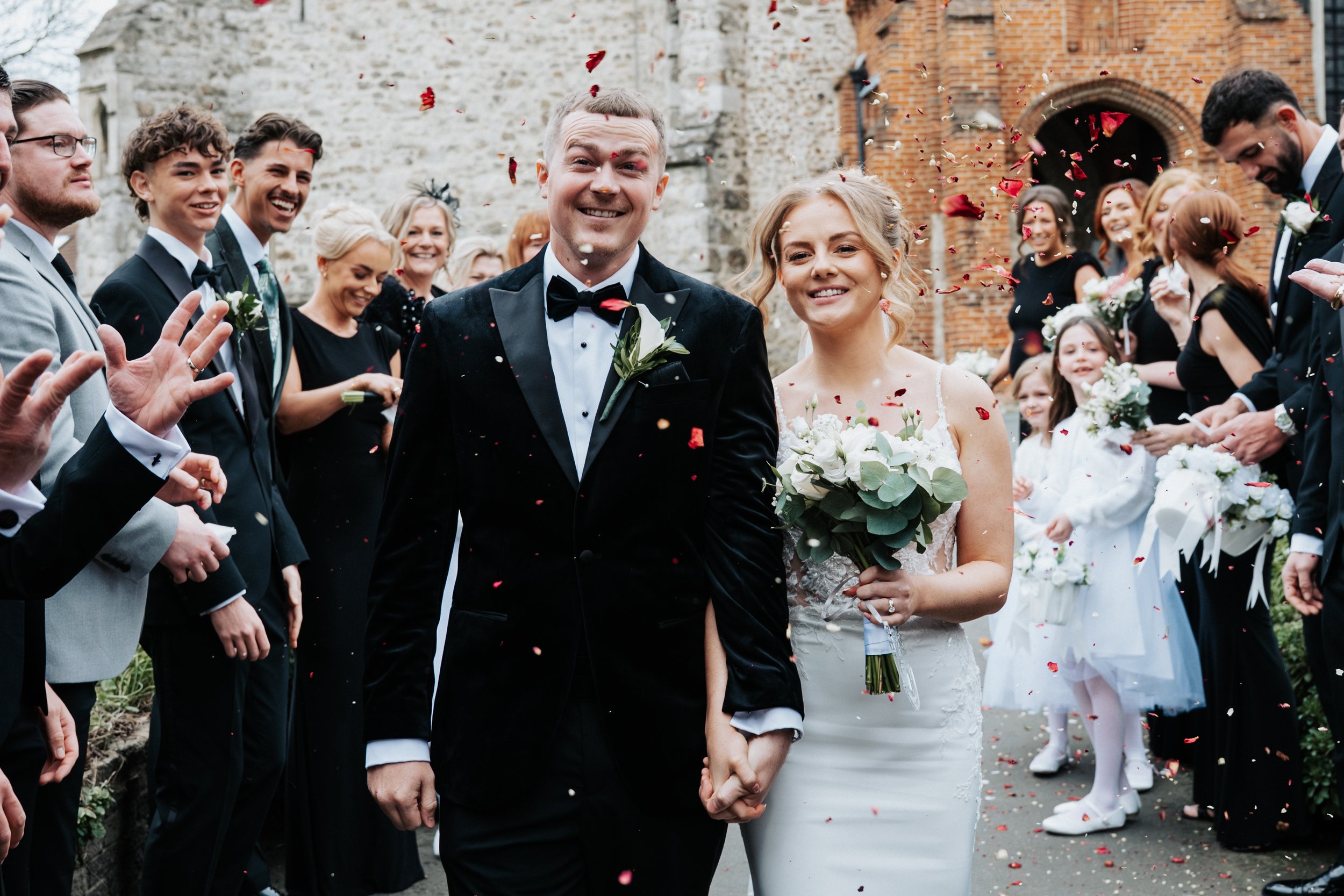 A newlywed couple walks hand-in-hand through a crowd throwing confetti outside of a brick building, with the bride holding a bouquet of white flowers and green foliage. The groom is wearing a black tuxedo with a white shirt and a black bow tie, and t