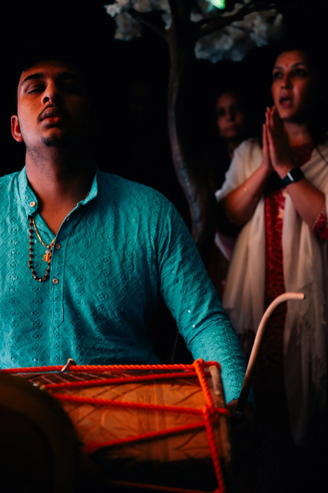 A man in a blue traditional outfit playing a drum, with two women in the background performing a prayer or greeting with folded hands, under a tree.