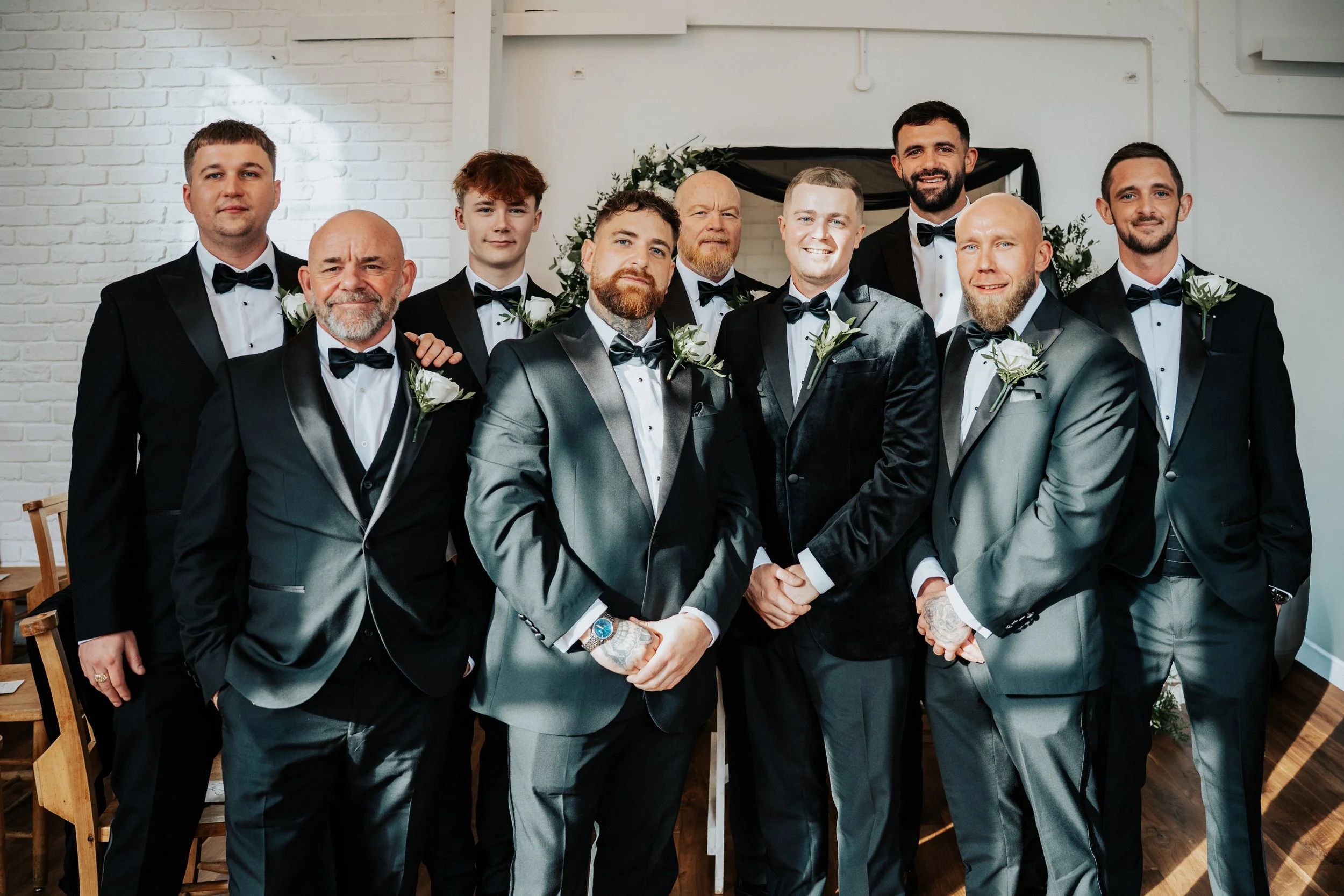A group of eleven men dressed in formal black, gray, and silver tuxedos with bow ties, standing together indoors for a wedding photo. Eight of them are wearing white shirts and three are wearing darker shirts, all with boutonnières on their lapels. T