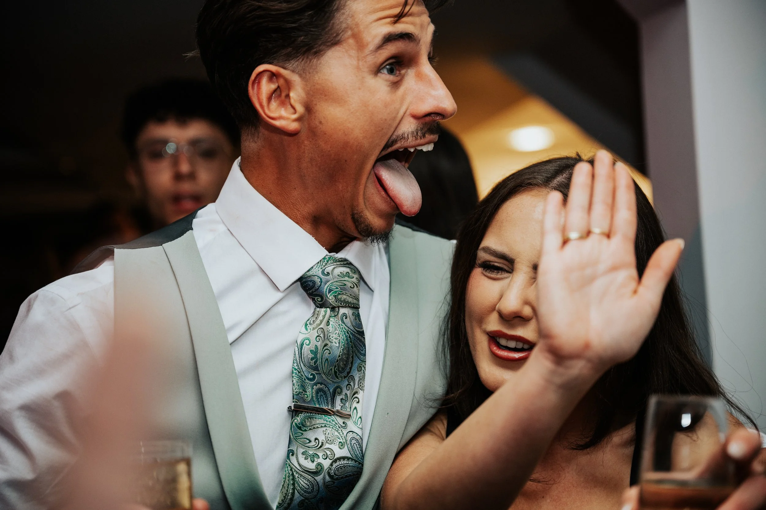 A man in a suit and tie with an open mouth and tongue sticking out, appearing excited or playful, standing close to a woman with long dark hair who is smiling and appears to be shielding her face with her hand, in a lively indoor setting.