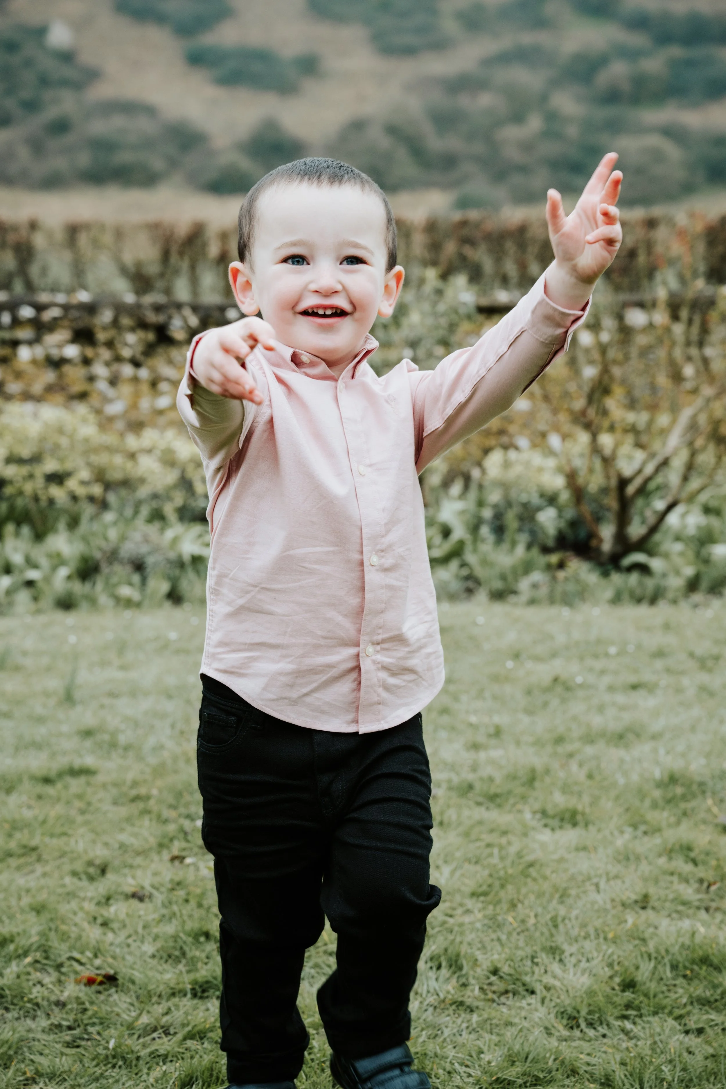 A young boy with short brown hair, wearing a light pink button-up shirt and black pants, is smiling and reaching out with both hands, standing outdoors on a grassy field with a stone wall and hills in the background.
