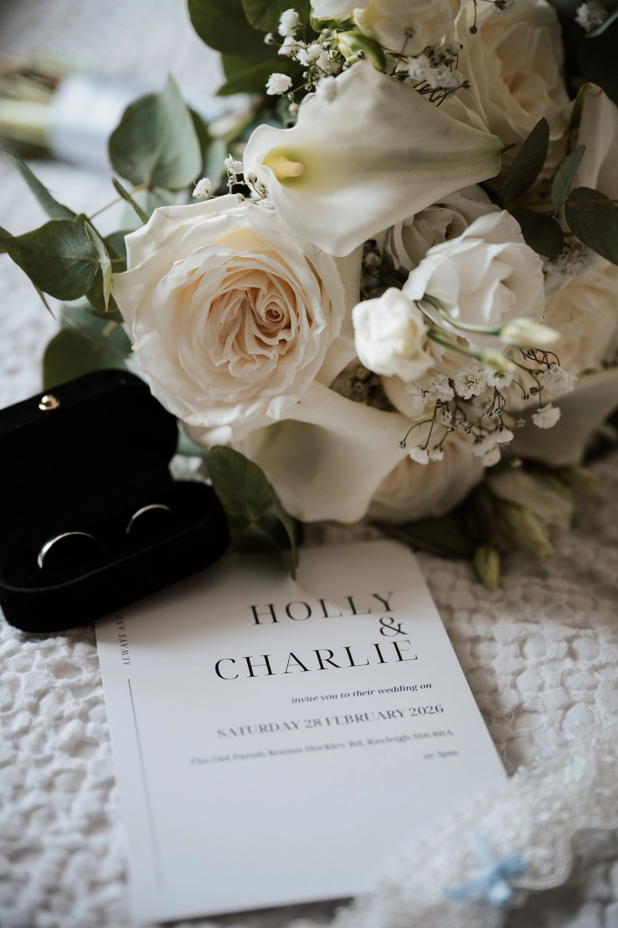 A wedding invitation for Holly and Charlie, a bouquet of white roses, calla lilies, and greenery, a black ring box with silver rings, and a white lace fabric background.