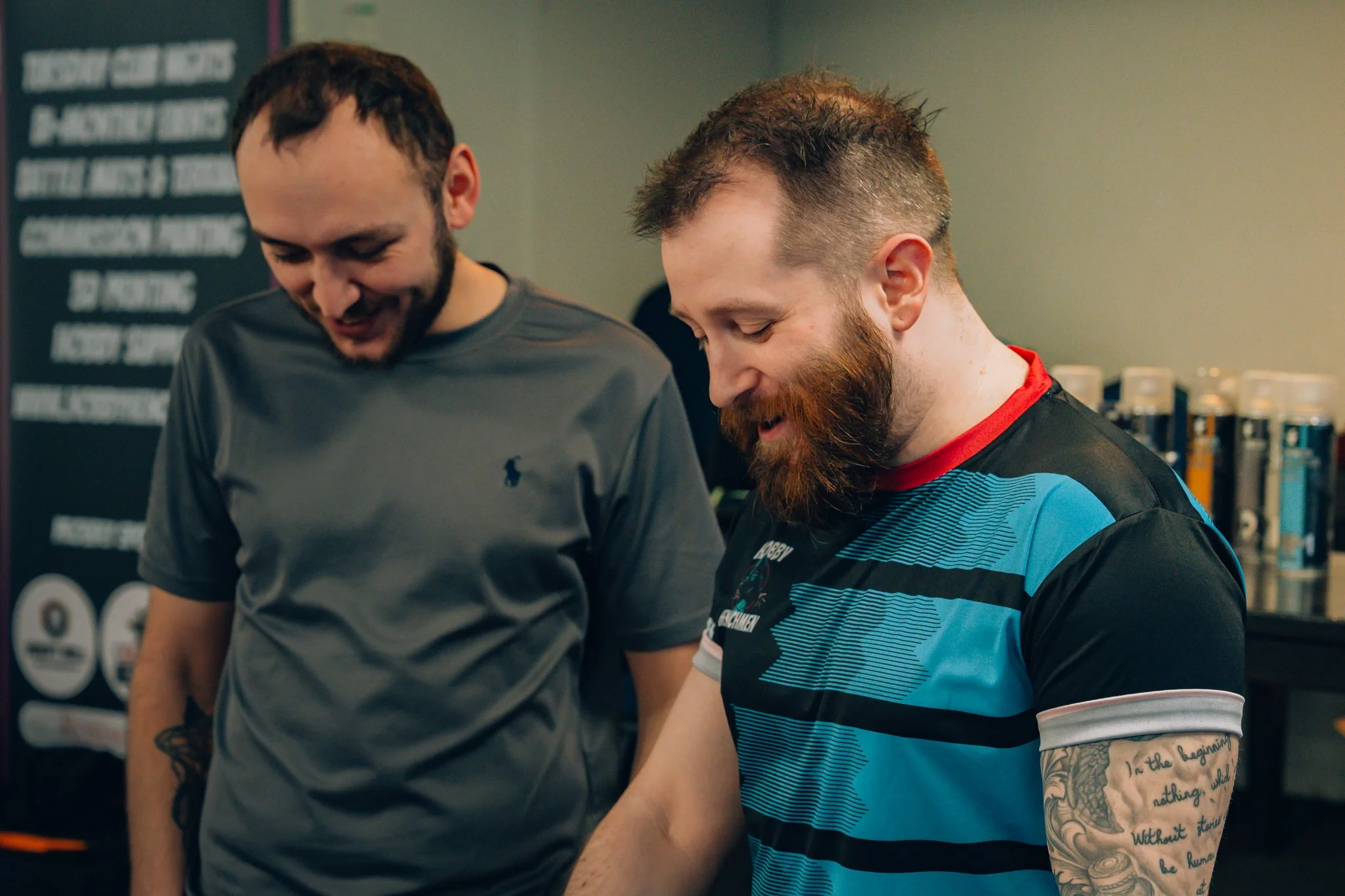 Two men smiling and looking down, standing indoors with a dark background and shelves with cans, brewers, or spices.
