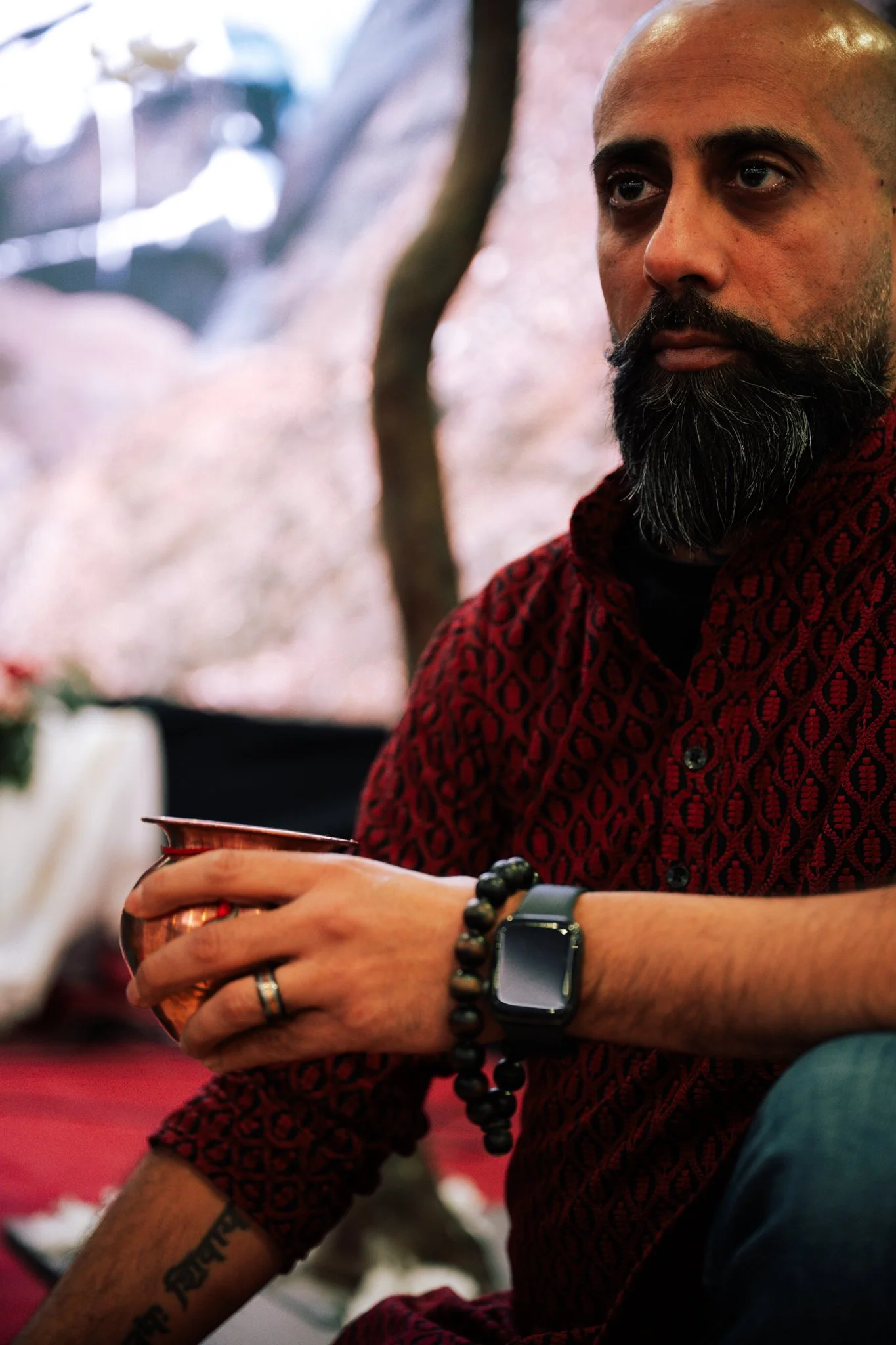 A man with a beard and shaved head wearing a red patterned shirt, holding a small copper vessel in his hand, with a smartwatch and a beaded bracelet on his wrist. The background is blurred with outdoor scenery.