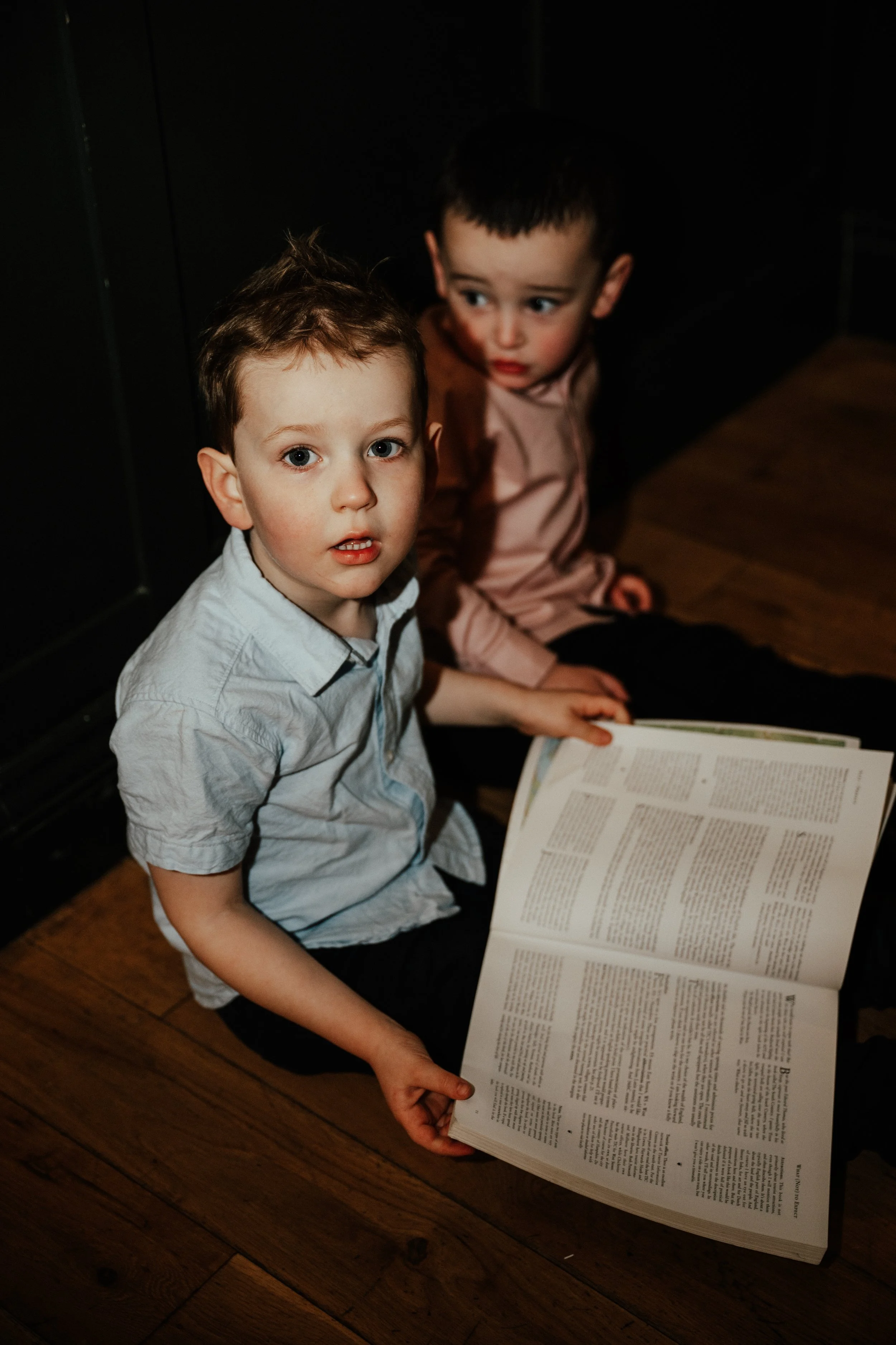 Two young boys sitting on a wooden floor, one holding an open book, with a dark background.