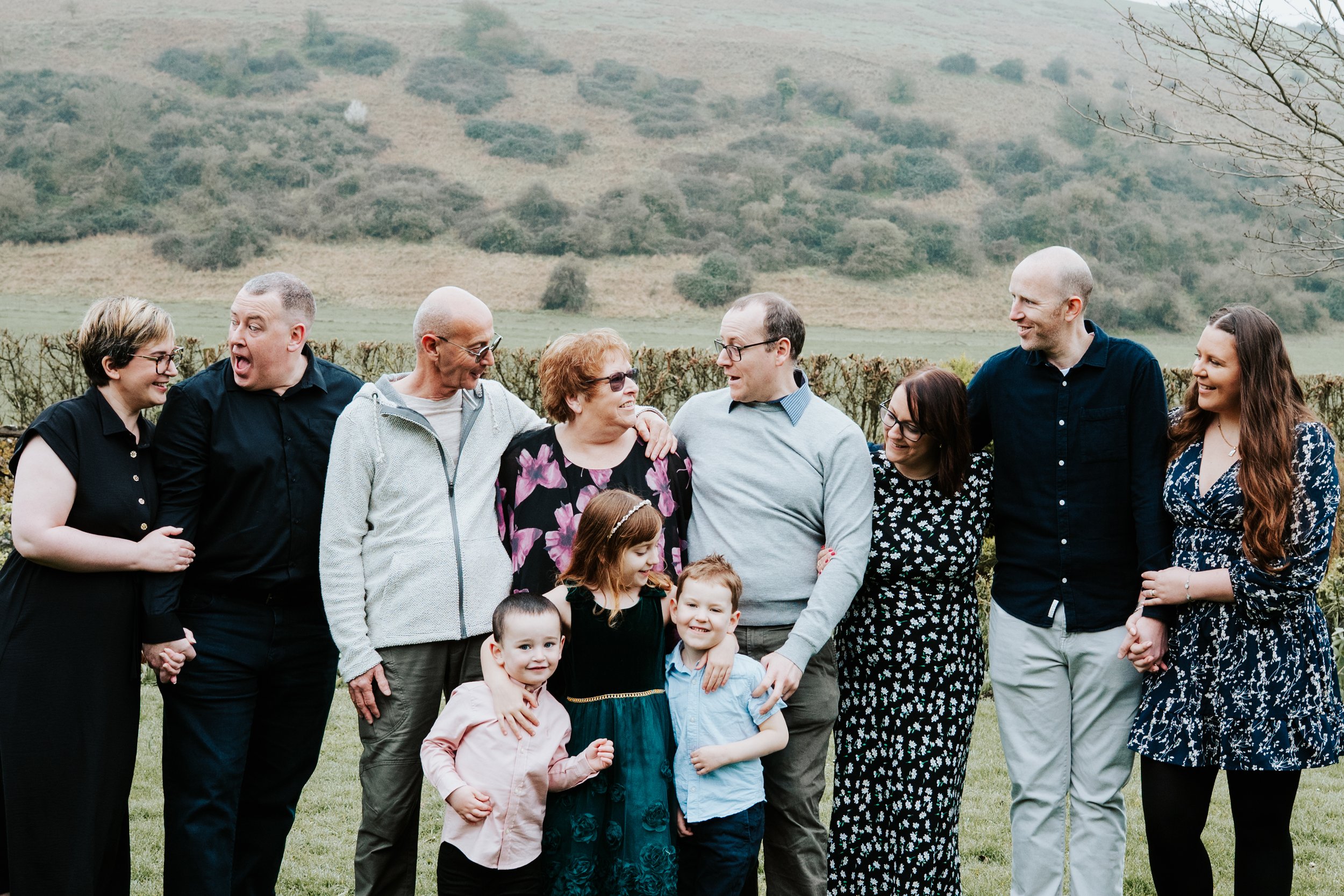 Group of people standing together outdoors on grass, holding hands and smiling, with a foggy hillside in the background.