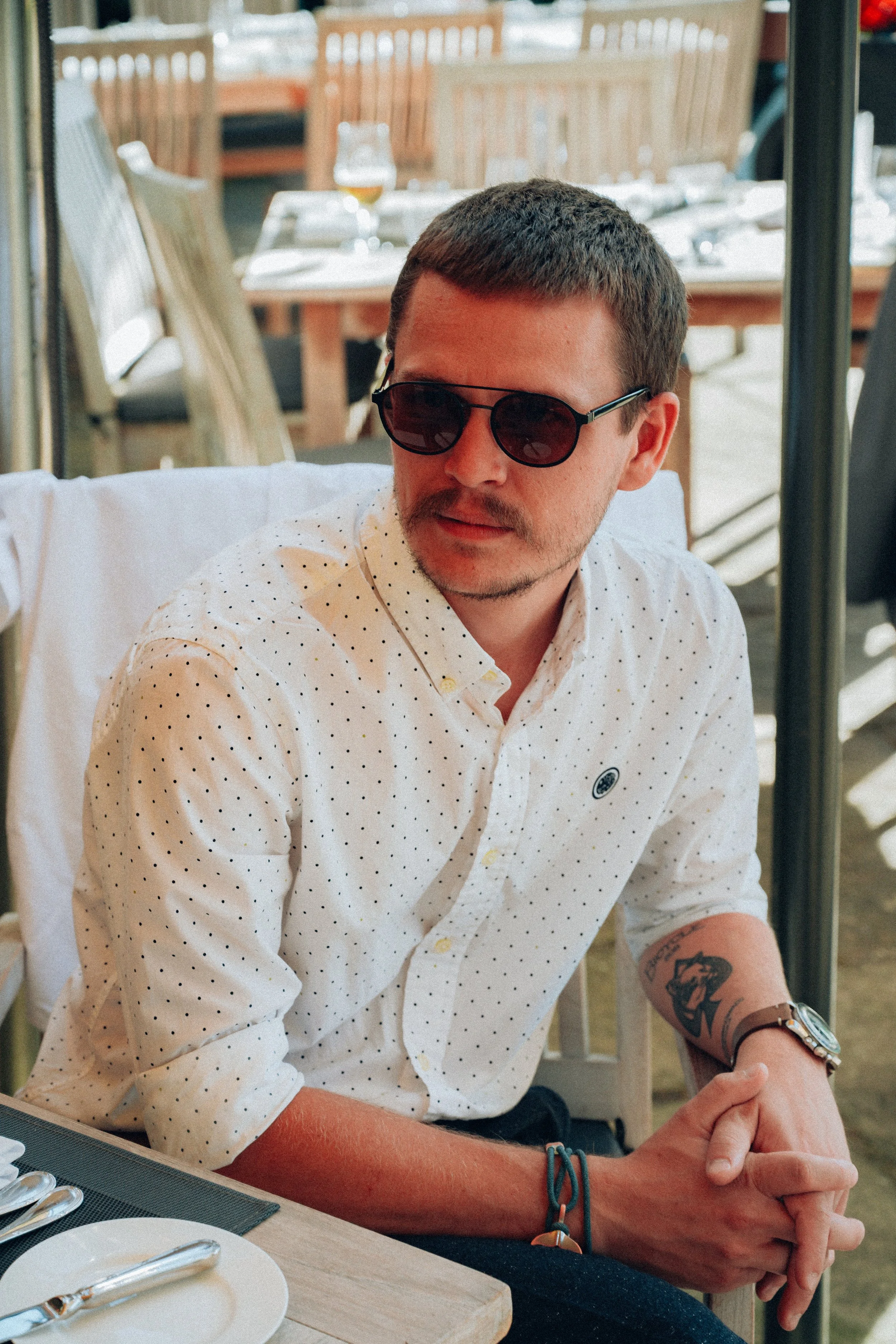 A man wearing black sunglasses and a white shirt with small black polka dots seated at a table in a restaurant or cafe.