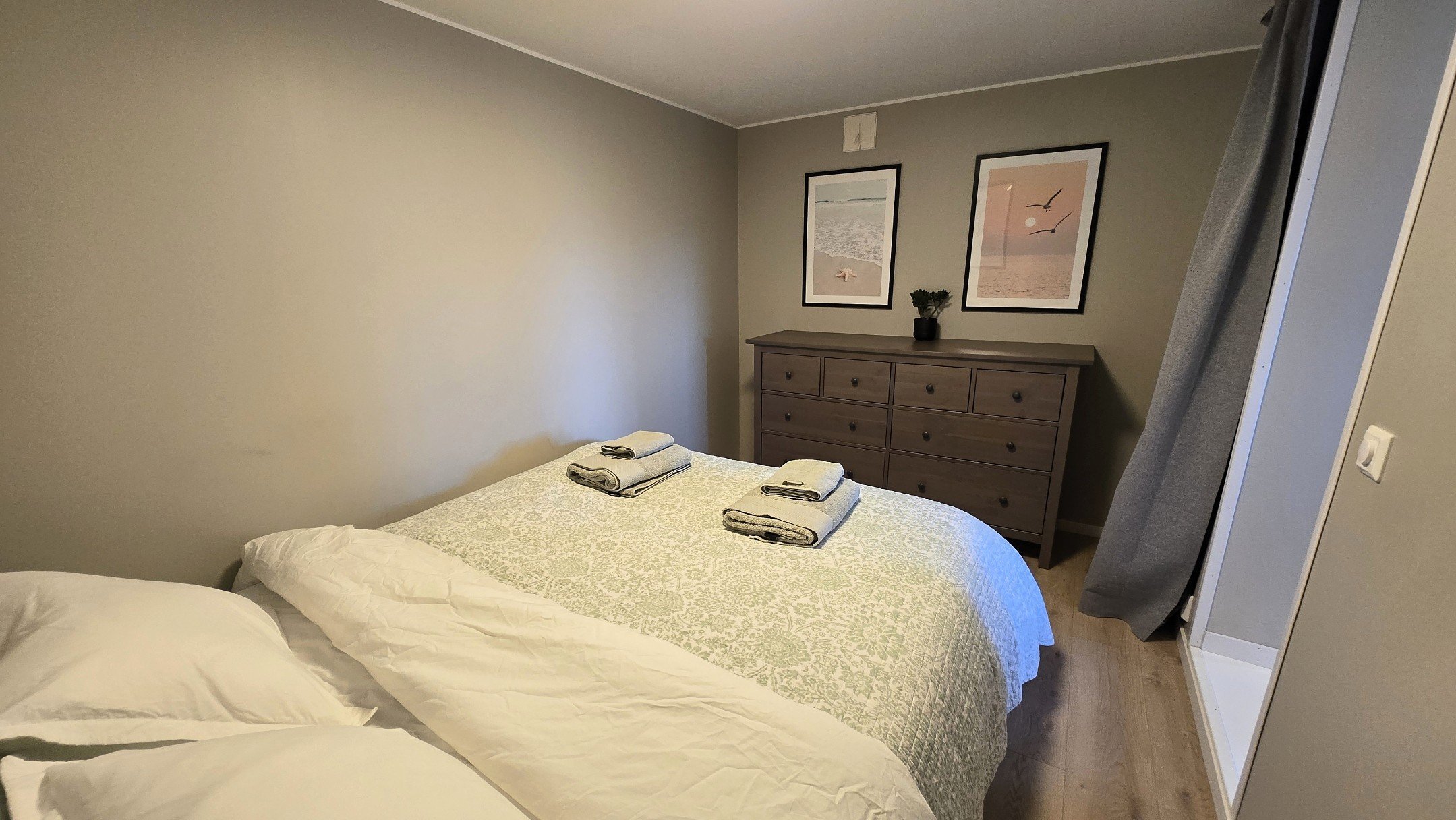 A bedroom with two beds, each having white pillows and white bedding, a gray dresser with nine drawers, two framed beach-themed pictures hanging above the dresser, and a small potted plant. Curtains hang to the side of a doorway, which opens to a sho